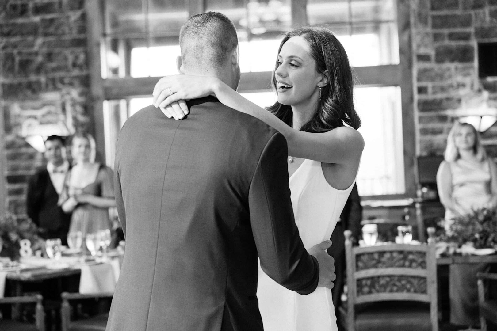 A couple is dancing together in a warmly lit room at The Ridge at Back Brook, with guests seated and standing in the background, celebrating their New Jersey wedding.