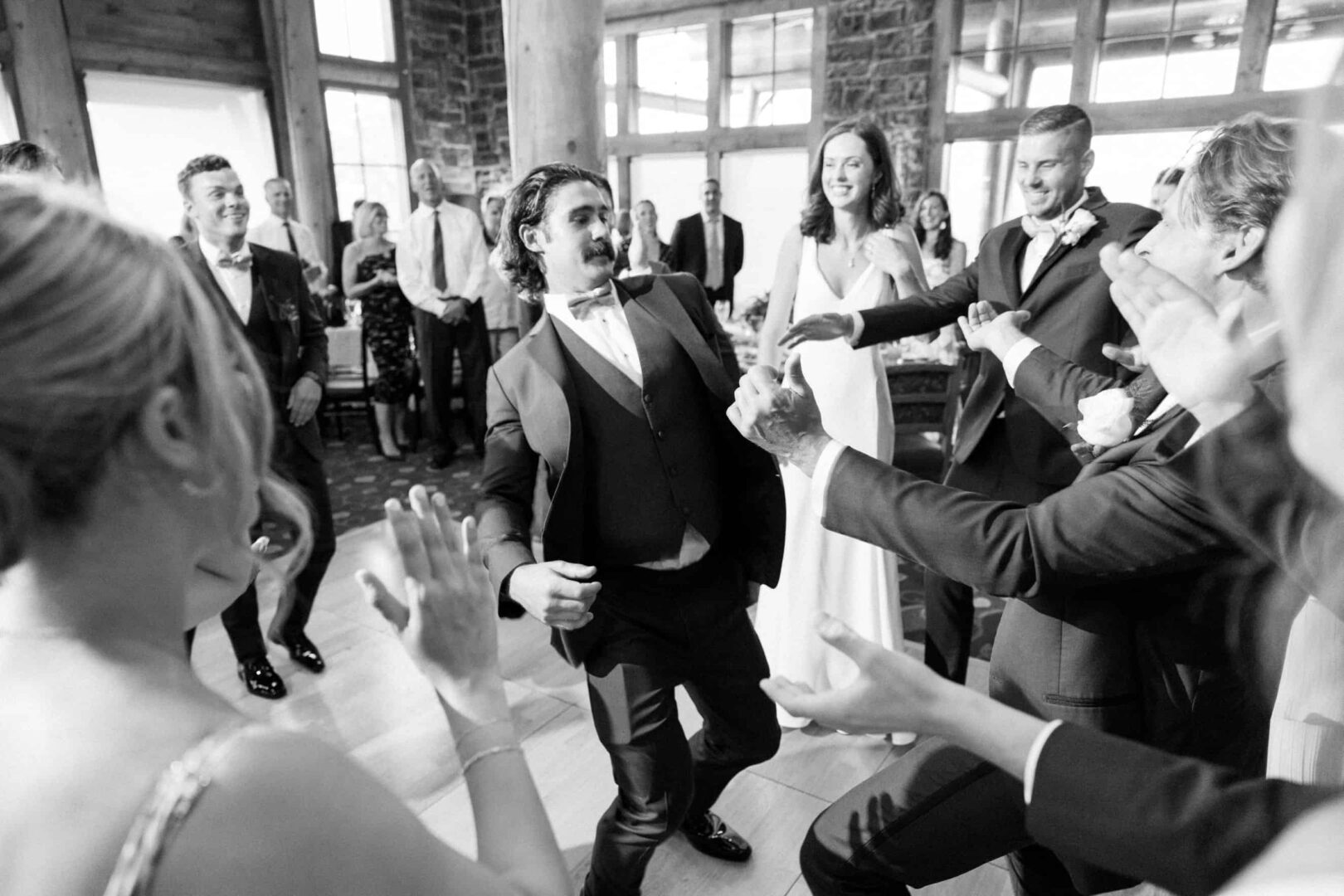 A group of people in formal attire dance energetically at an indoor event at The Ridge at Back Brook, a prestigious country club in New Jersey, surrounded by onlookers clapping and cheering.