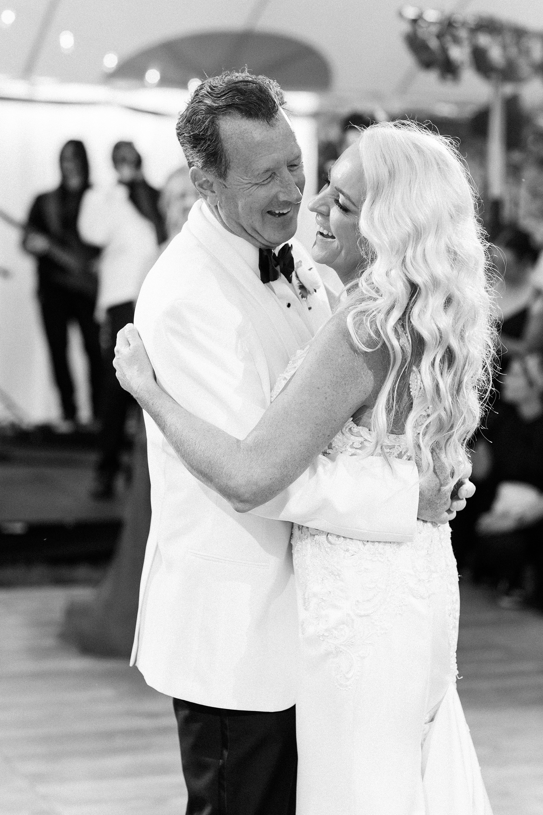 A couple dressed in white formal attire dances closely together at an event while a band plays in the background.