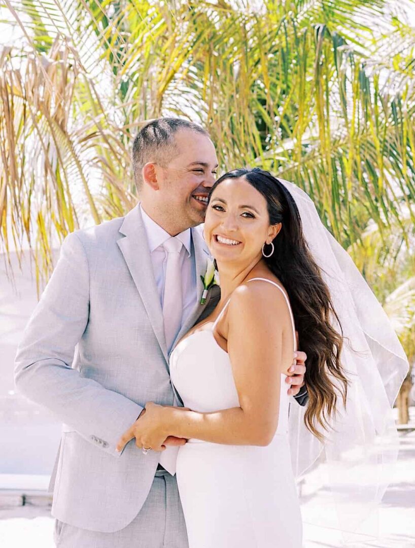 A smiling couple in wedding attire stands close together under palm trees on a sunny day at their beach wedding in Wildwood, New Jersey. The man, in a light gray suit, hugs the woman, who is wearing a white dress and veil.