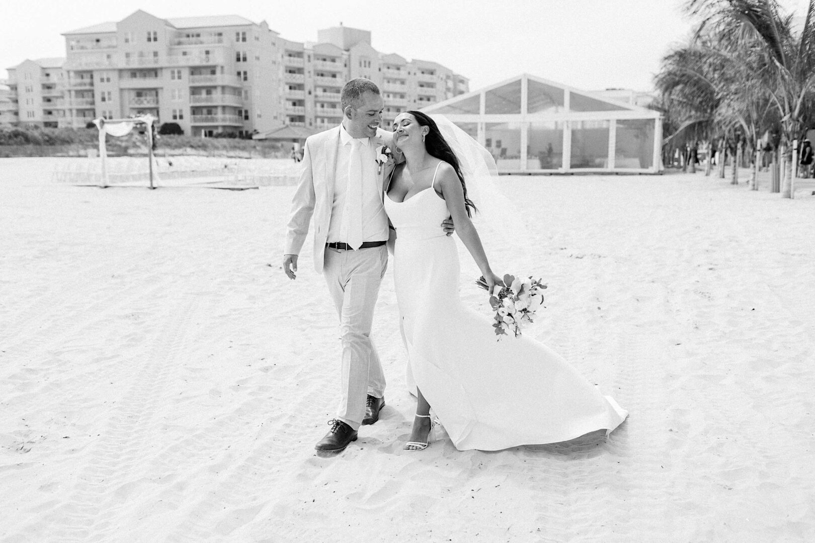 A bride and groom walk arm in arm on a sandy Wildwood, New Jersey beach. Both are dressed in white attire; she holds a bouquet. Buildings and a structure are visible in the background. The scene is in black and white.