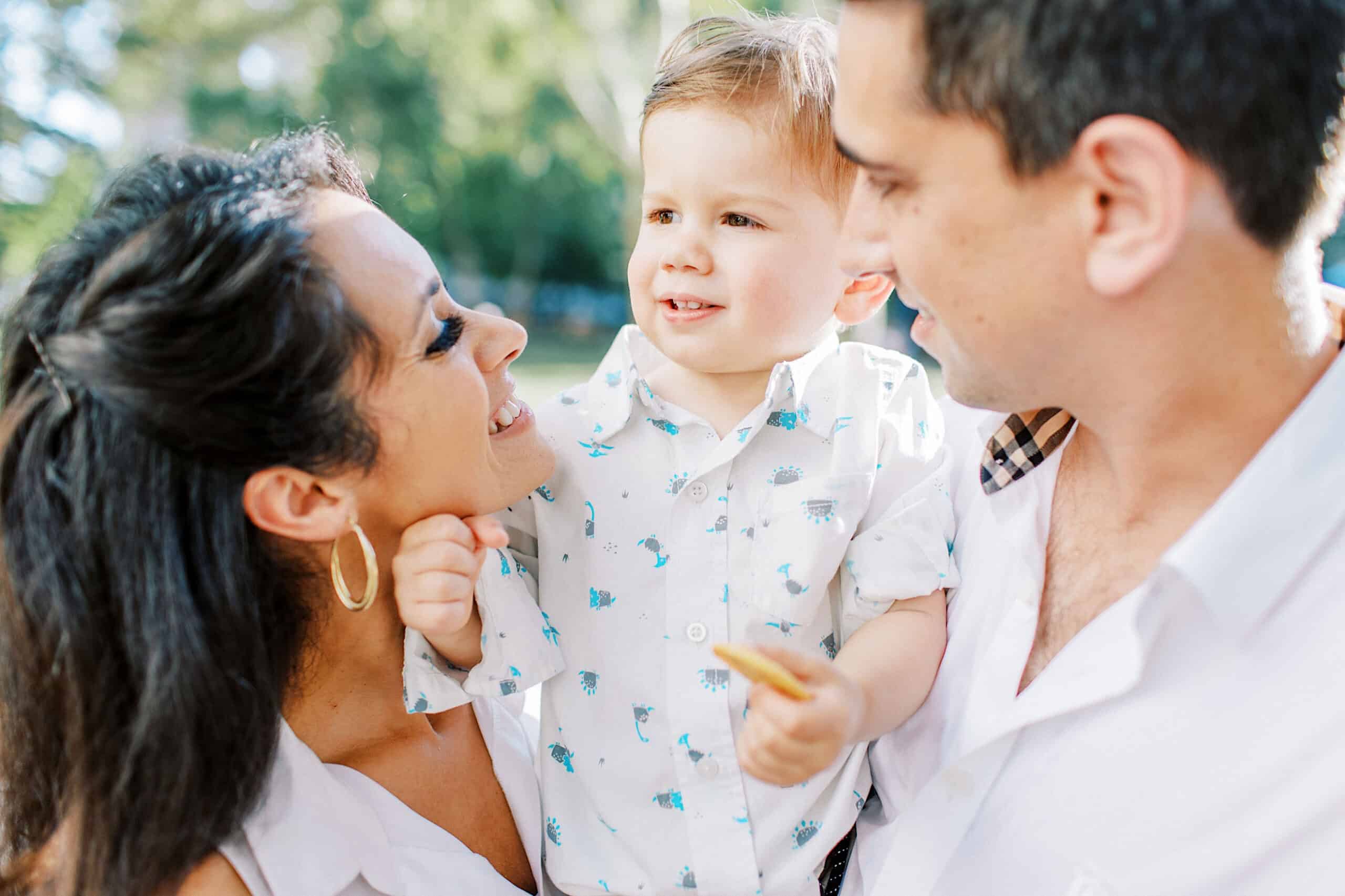 A woman and man smile at a young boy they are holding between them outdoors in matching white shirts during family photos at Rittenhouse Square.