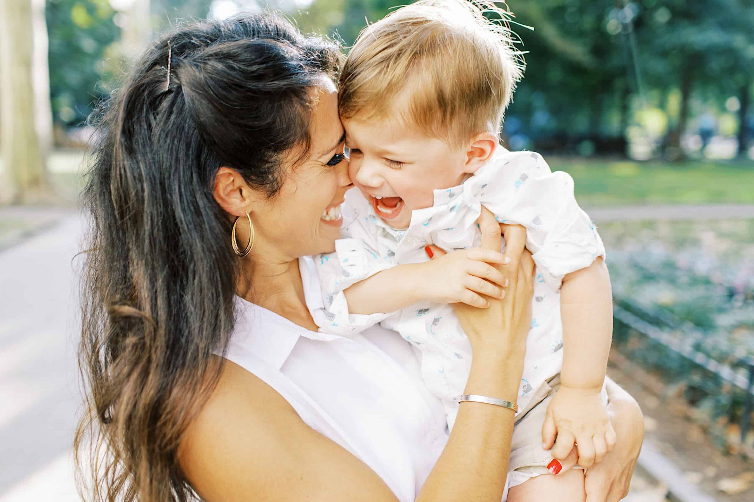 A woman holds and smiles at a young child outdoors in a park, both appearing happy and engaged—a beautiful moment perfect for family photos at Rittenhouse Square.