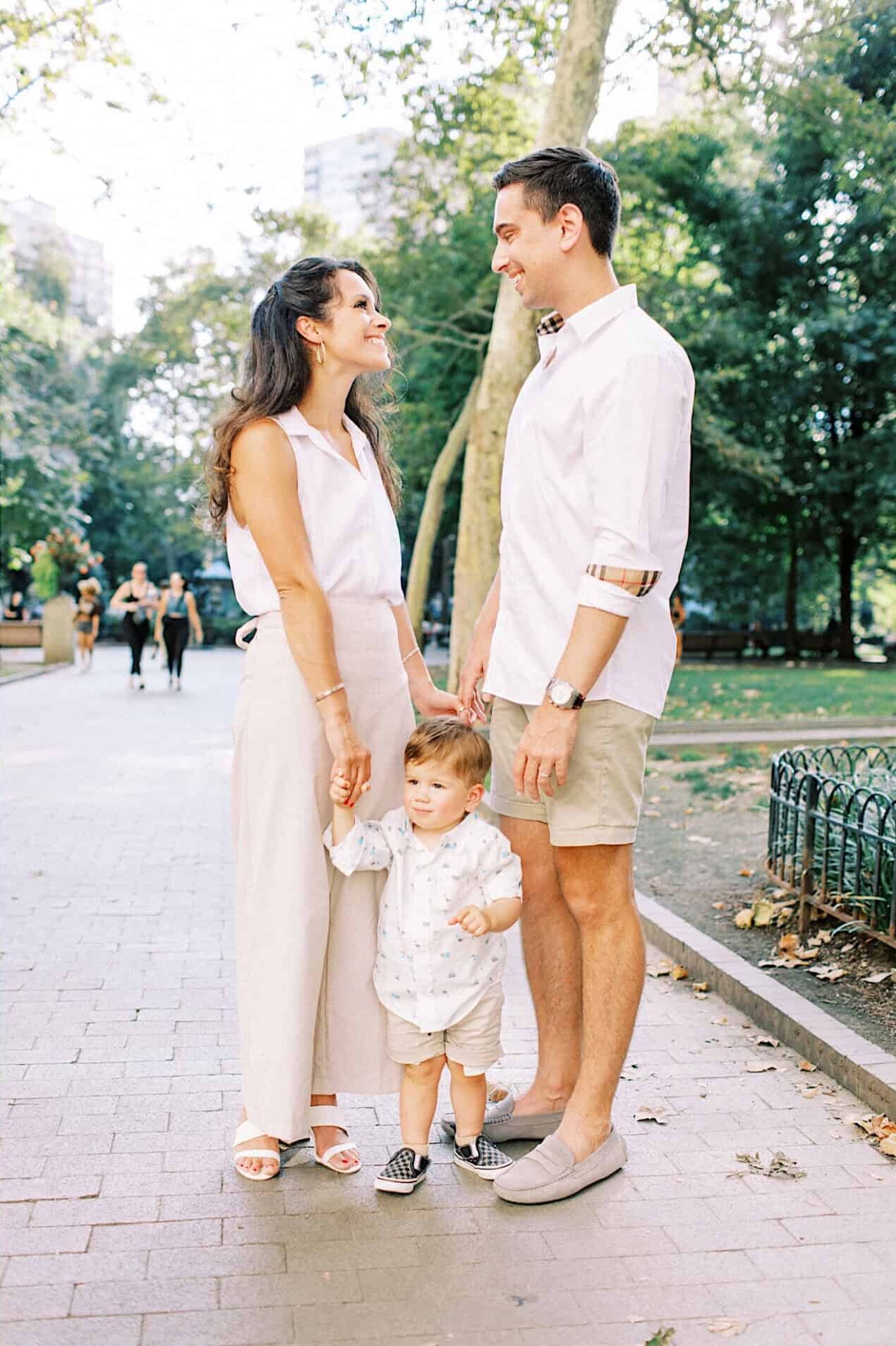 A family of three with a young child stands and interacts on a tree-lined path in Rittenhouse Square, capturing family photos on a sunny day.
