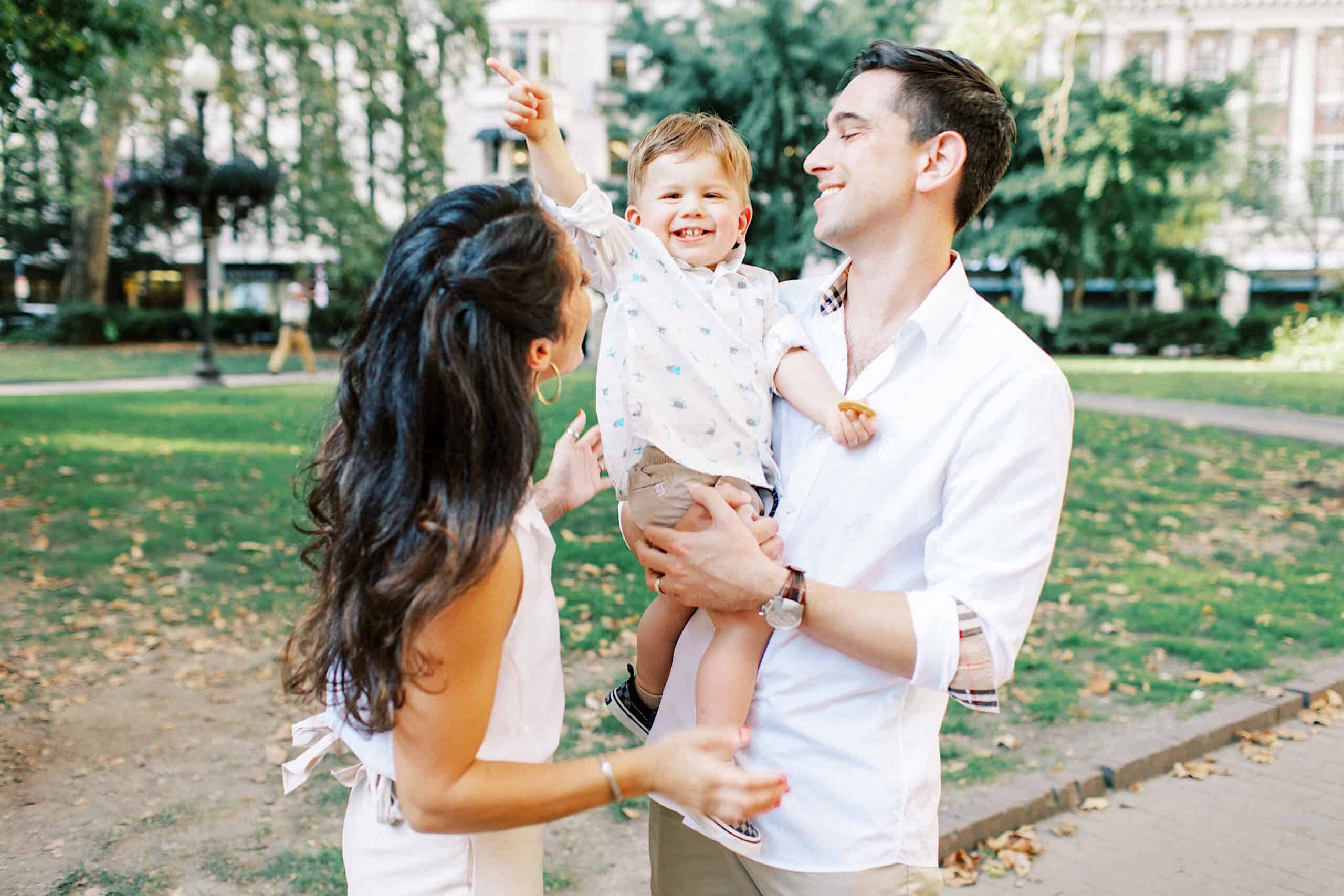 A man holds a smiling young boy while a woman stands beside them in a park with green trees and buildings in the background, capturing family photos at Rittenhouse Square.