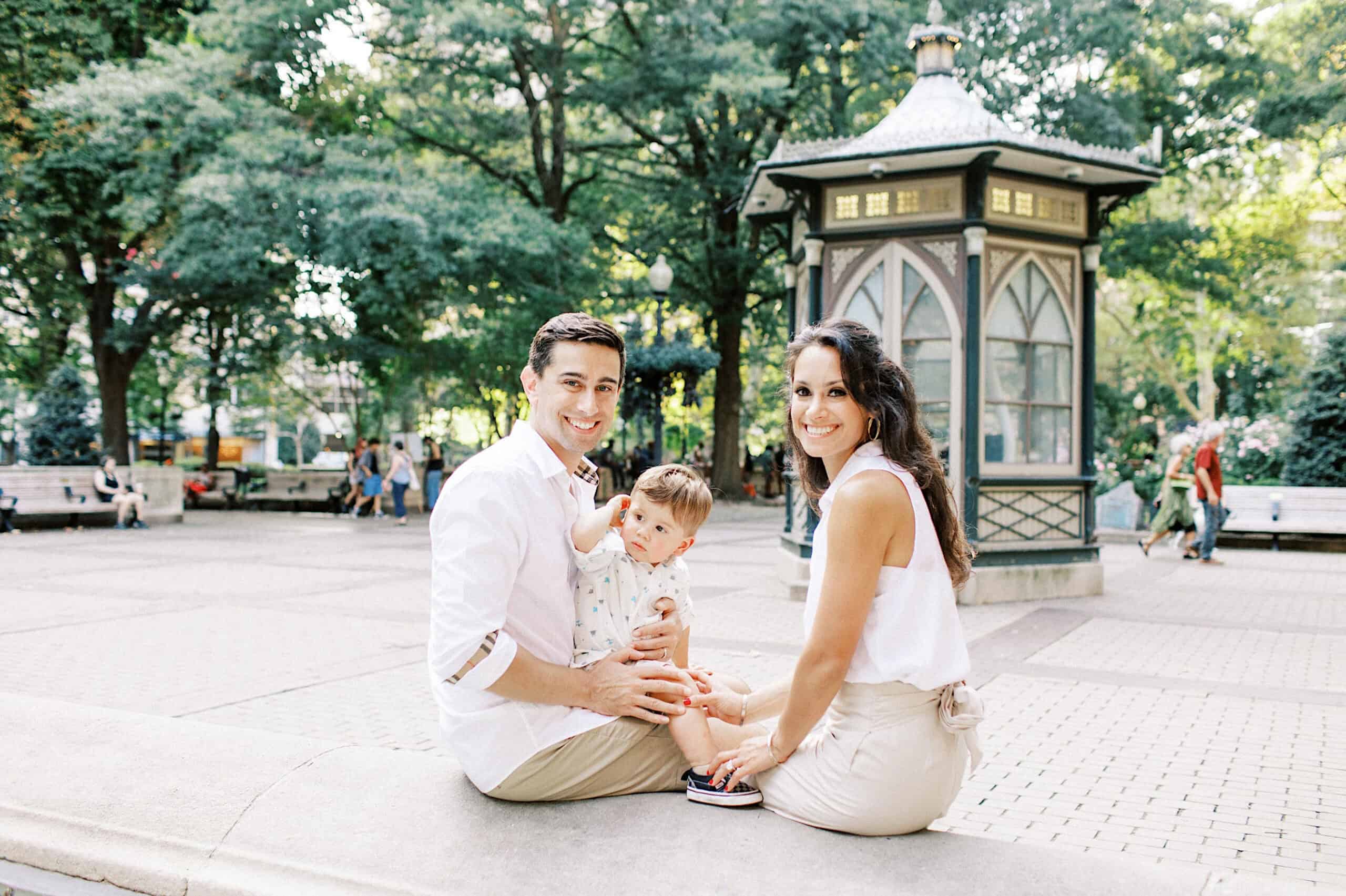A man, woman, and young child sit together on a concrete ledge in a park near a small pavilion, capturing family photos at Rittenhouse Square with trees and benches in the background.
