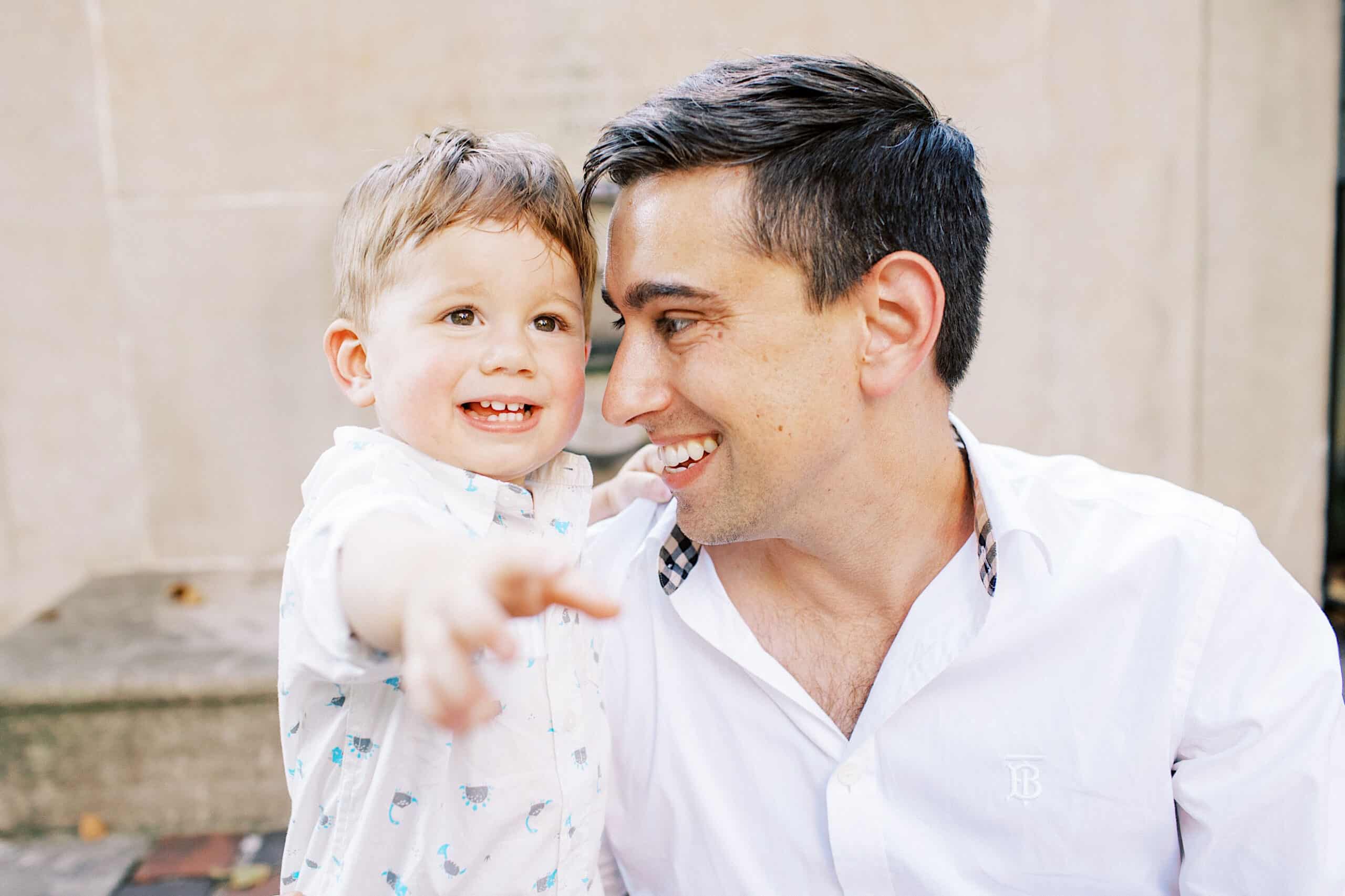 A man in a white shirt sits closely with a smiling young boy pointing forward. Both appear happy and are looking in the same direction, capturing the warmth of family photos at Rittenhouse Square.