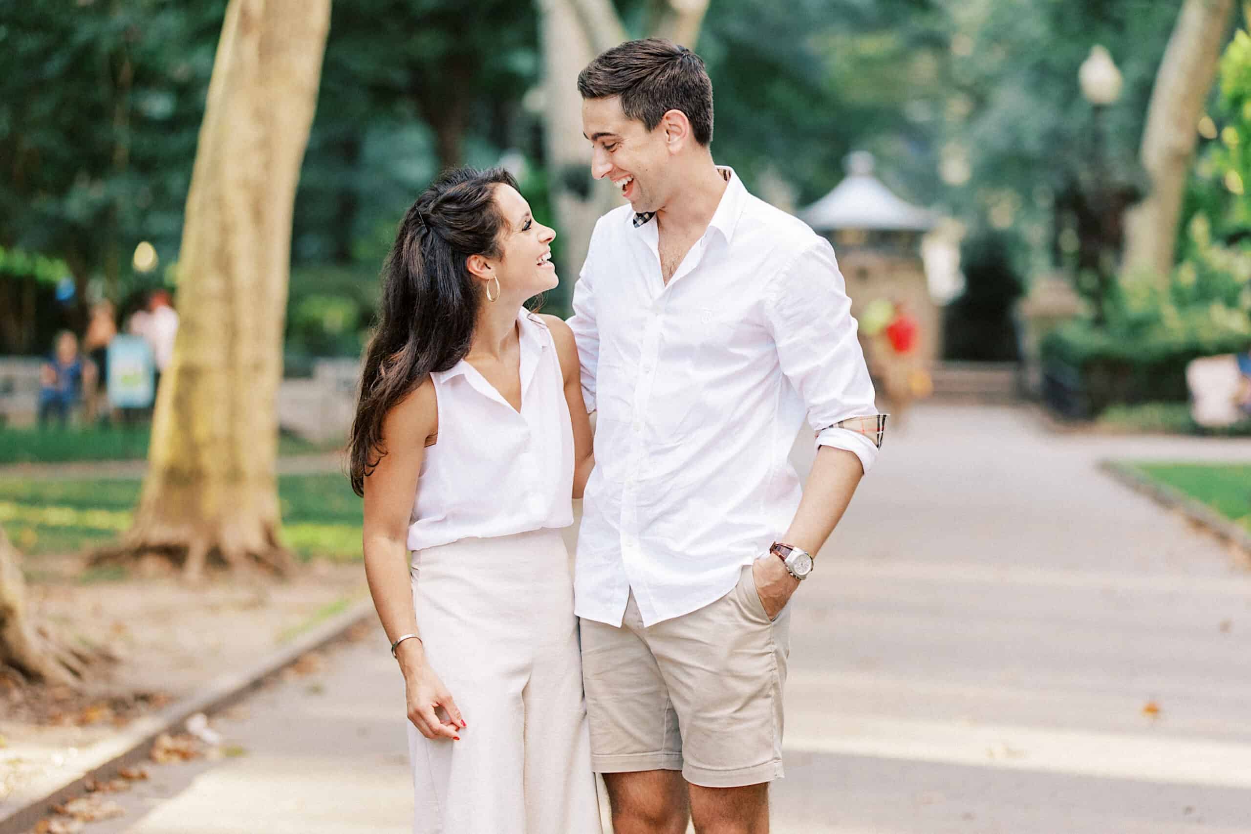 A man and a woman stand on a park path, smiling at each other in light summer clothes. Trees and people fill the background, capturing the warmth of family photos at Rittenhouse Square.