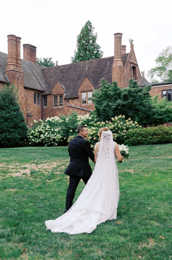 A bride and groom stand on a lawn in front of a large brick mansion, surrounded by trees and greenery.