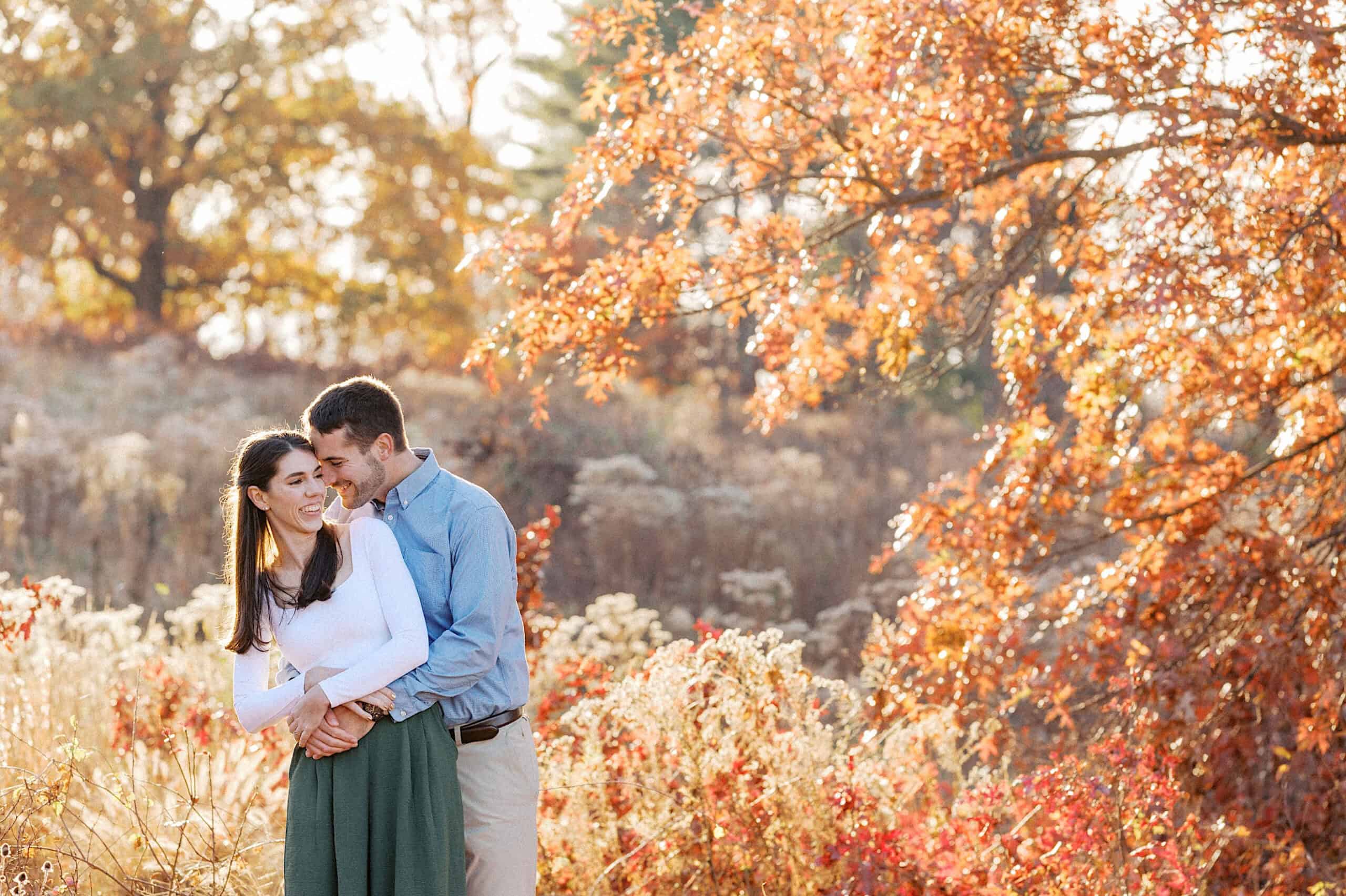 A man and woman stand close together, smiling, in a sunlit field with autumn trees and foliage—a perfect moment for golden hour fall engagement photos.