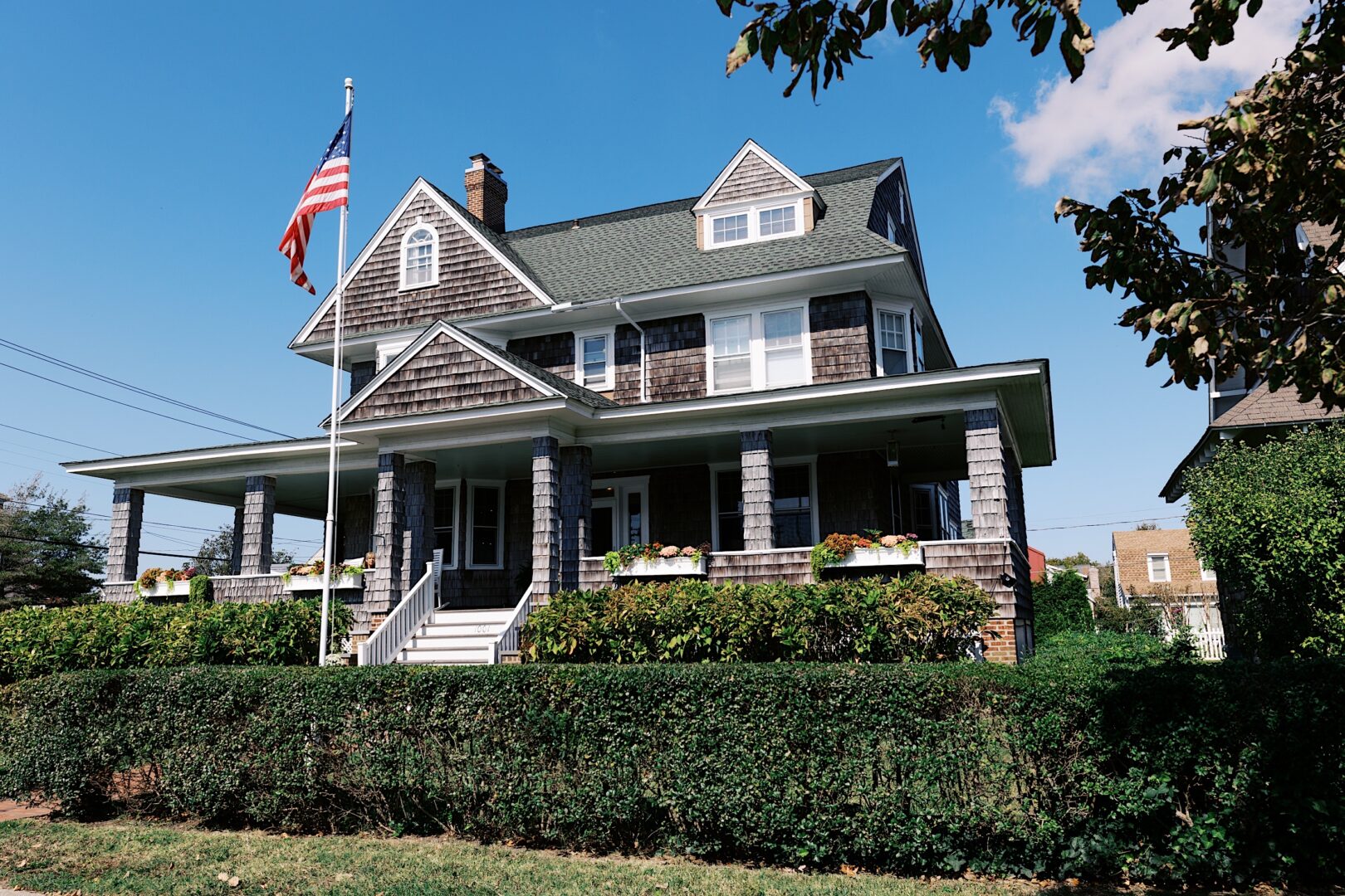 A large, two-story house with a porch and shingle exterior, an American flag on a pole, and neatly trimmed hedges in the front yard exudes charm reminiscent of a Corinthian Yacht Club of Cape May wedding.