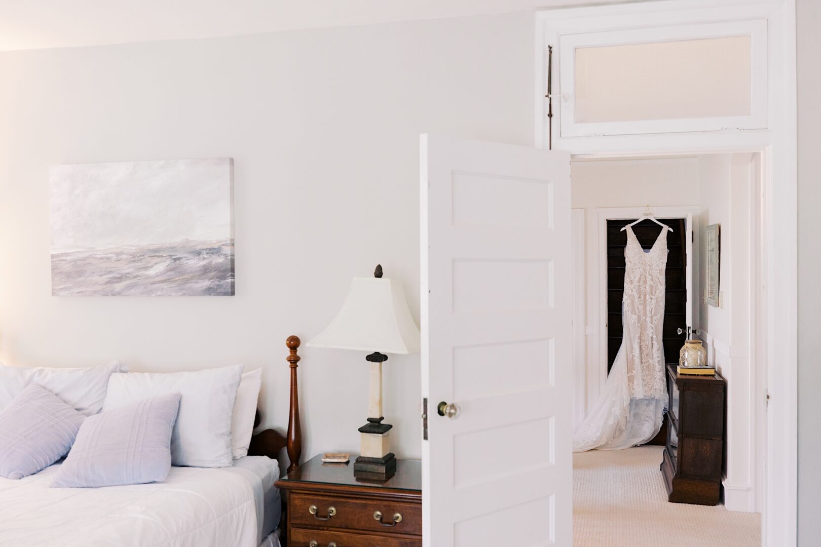 A bedroom with a bed, nightstand, and lamp is shown. A doorway opens to a room where a wedding dress, reminiscent of a Corinthian Yacht Club of Cape May Wedding, hangs elegantly on the wardrobe.