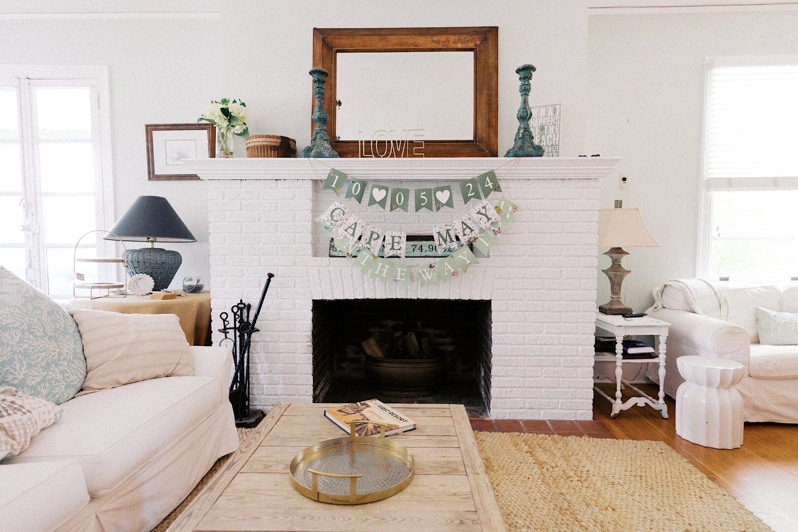 A cozy living room with a white brick fireplace adorned with a banner displaying "Cape May, the way I am" and a date, "10-05-24," reminiscent of a Corinthian Yacht Club of Cape May wedding. The space features a beige couch, lamps, and tasteful decor items.