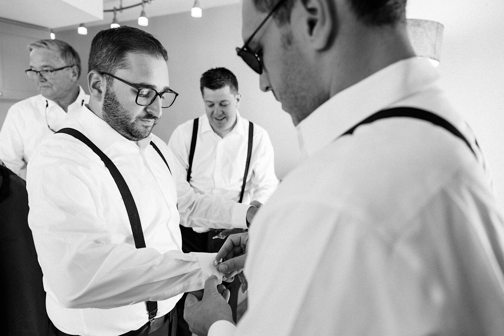 A group of men in white shirts and suspenders are getting ready at the Corinthian Yacht Club of Cape May Wedding, with one helping another adjust his sleeve. They appear focused and are indoors.