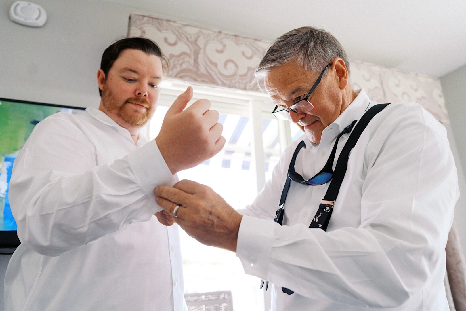 Two men in white shirts adjusting their cuffs stand in a room with a window and patterned curtain. One, wearing glasses and suspenders, prepares for the Corinthian Yacht Club of Cape May wedding.