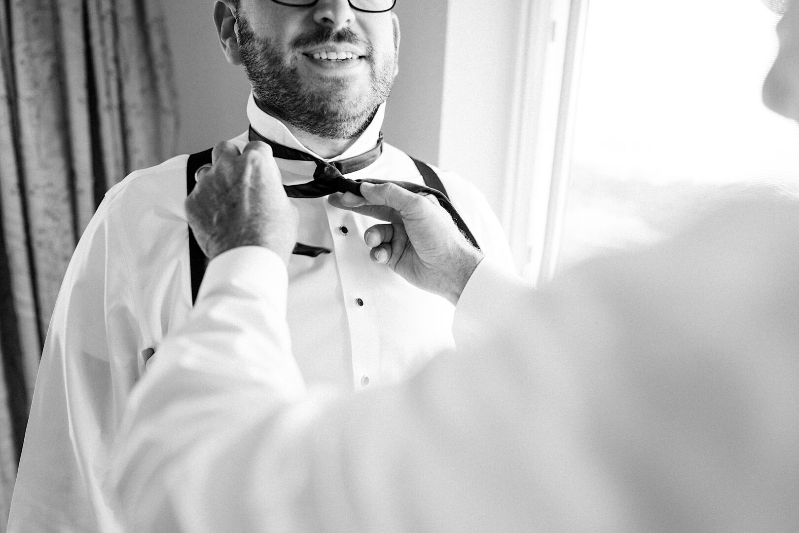 A person adjusts the bow tie of another, dressed in a white shirt and suspenders, near a window at the Corinthian Yacht Club of Cape May Wedding.