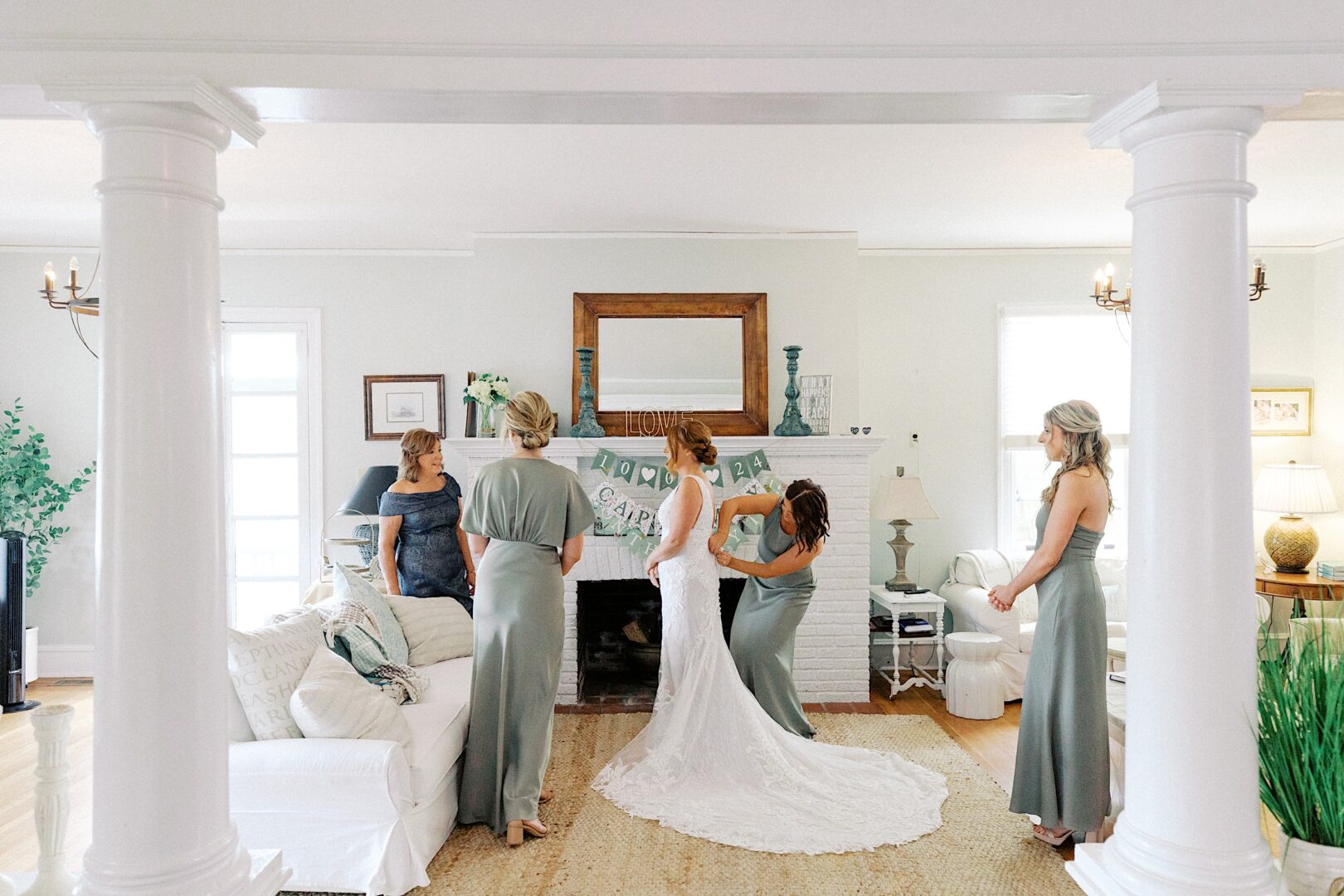 Bridal party prepares in a living room before the Corinthian Yacht Club of Cape May wedding. The bride, in a white gown, is surrounded by four women in matching green dresses. The room features a white sofa, a banner, and elegant pillar decor.