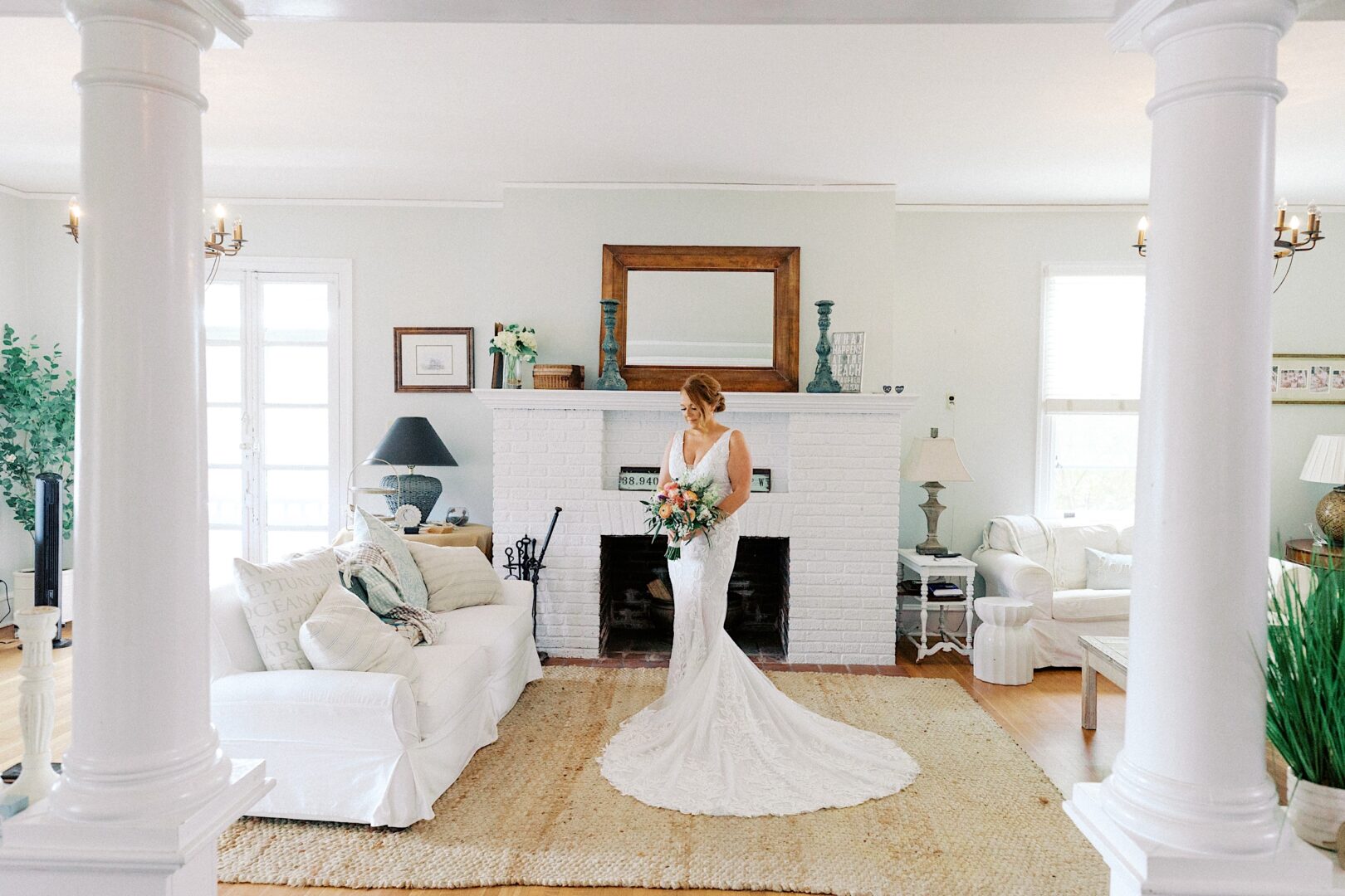 In a bright, elegant living room adorned with white couches, a fireplace, and decorative mirrors, the bride in her white dress radiates joy—capturing the timeless charm reminiscent of a Corinthian Yacht Club of Cape May wedding.