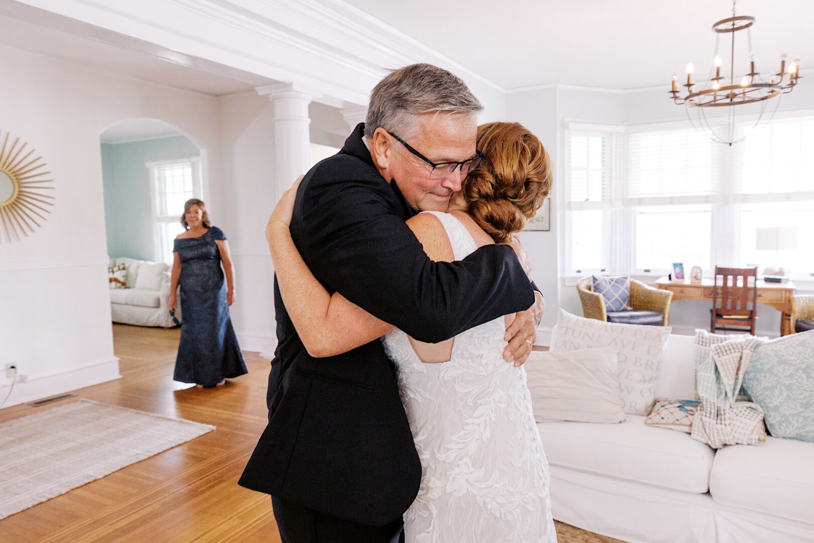 In a cozy living room, a person in a suit warmly embraces someone in a white dress, reminiscent of the joyful moments at a Corinthian Yacht Club of Cape May wedding. In the background, another figure stands gracefully in formal attire.