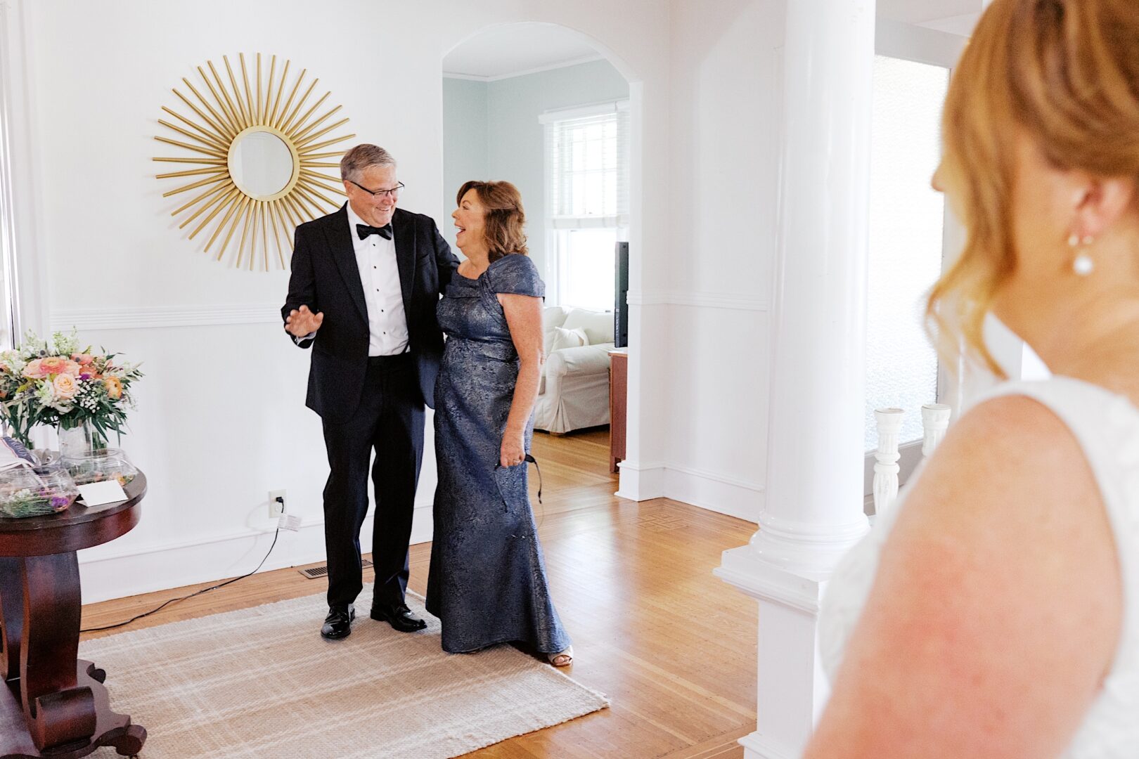 In a bright room adorned with a sunburst mirror, a couple in formal attire shares a smile at their Corinthian Yacht Club of Cape May wedding, while someone stands in the foreground.