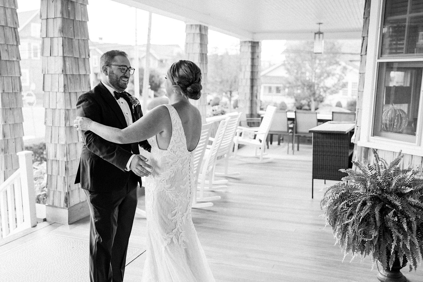 A bride and groom embrace on the spacious porch of the Corinthian Yacht Club of Cape May, both smiling amid rocking chairs and a table. The touching scene is beautifully captured in black and white.