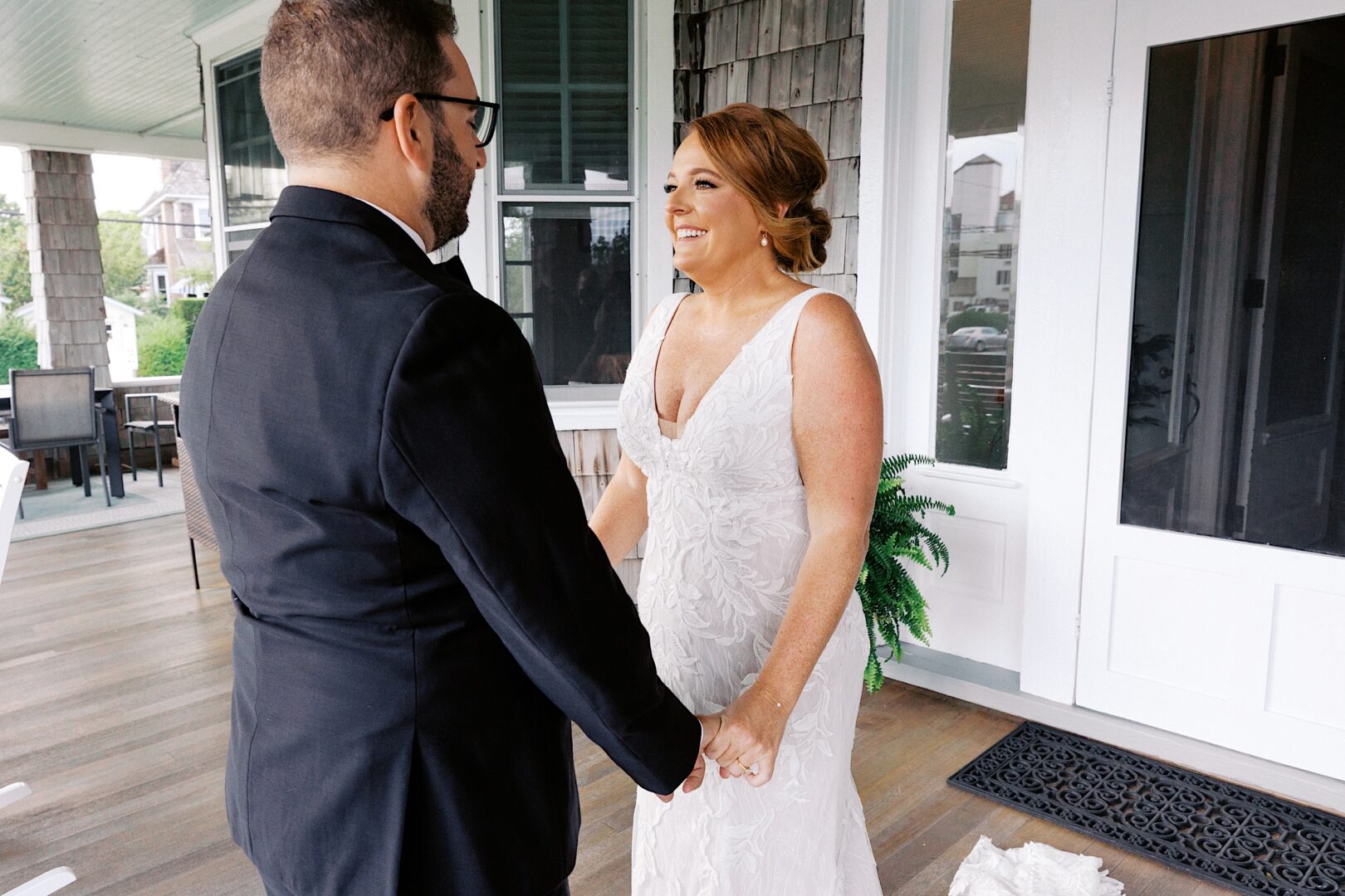 A couple stands together on the porch of the Corinthian Yacht Club of Cape May, holding hands and smiling in their elegant wedding attire.