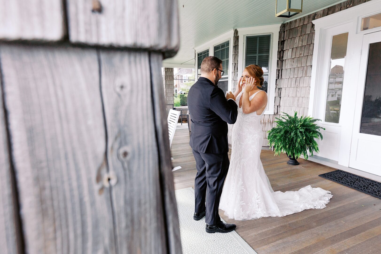 A bride and groom stand together on a wooden porch at the Corinthian Yacht Club of Cape May, with the bride covering her face emotionally and the groom gently touching her hands.