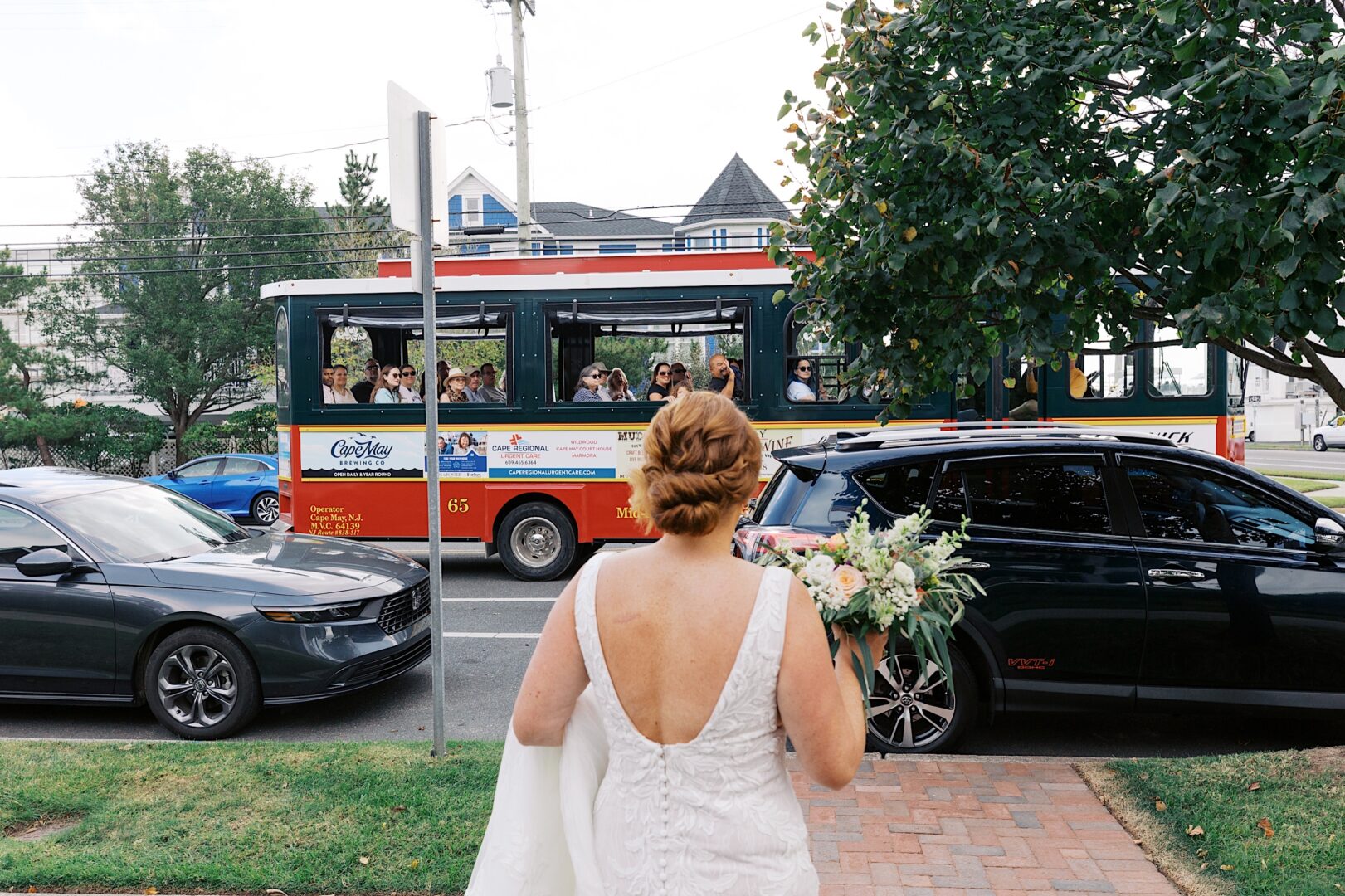 A bride in a white dress stands on the sidewalk, clutching her bouquet, as a trolley filled with dogs passes by, capturing the whimsical charm of a Corinthian Yacht Club of Cape May wedding.