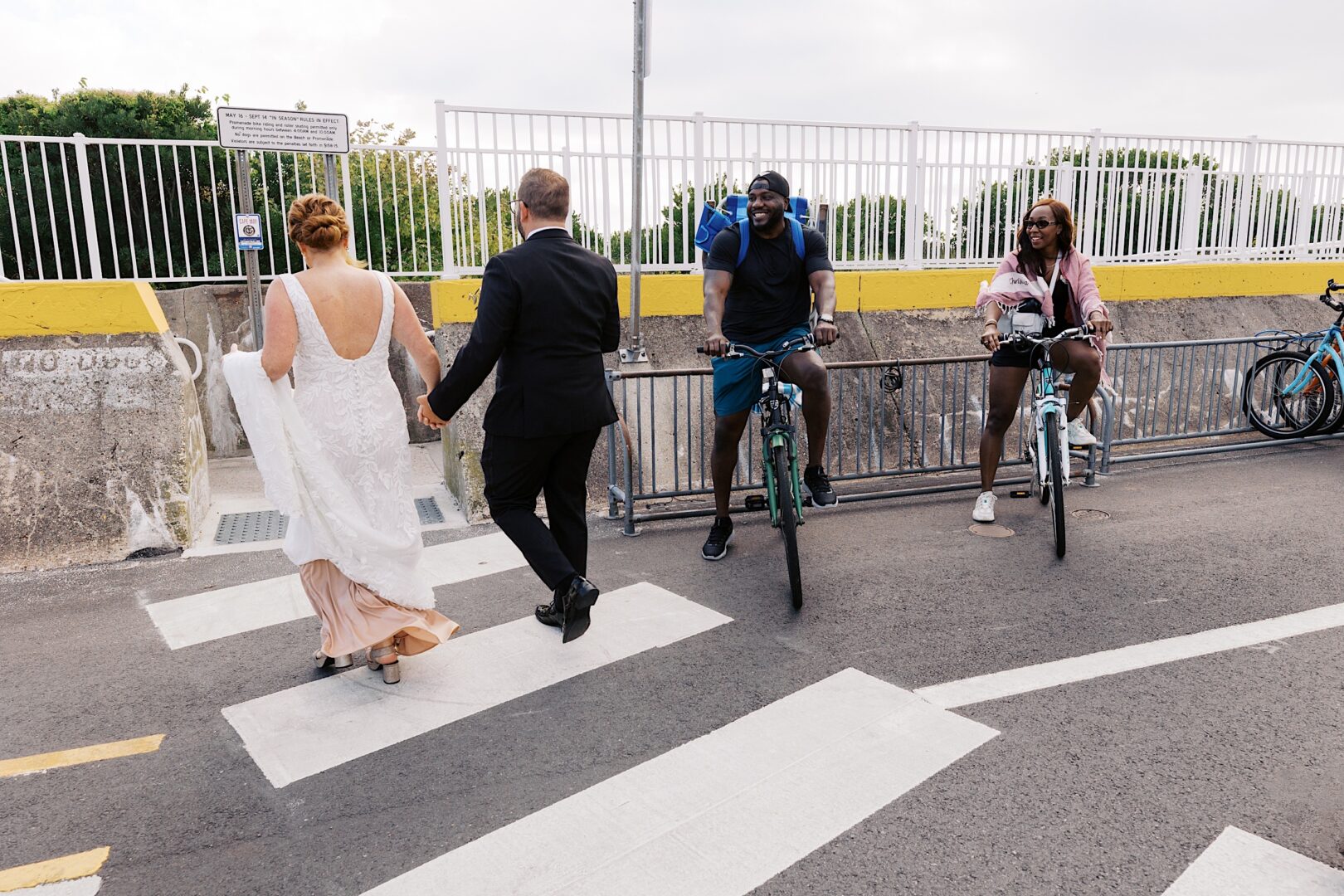 A couple in wedding attire walks on a crosswalk, capturing the essence of their Corinthian Yacht Club of Cape May wedding, as two cyclists look on, with a fence and trees in the background.