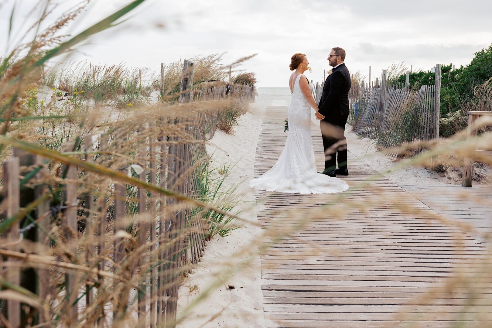 A bride and groom stand on a wooden walkway by the beach at the Corinthian Yacht Club of Cape May Wedding, surrounded by sand and grass.