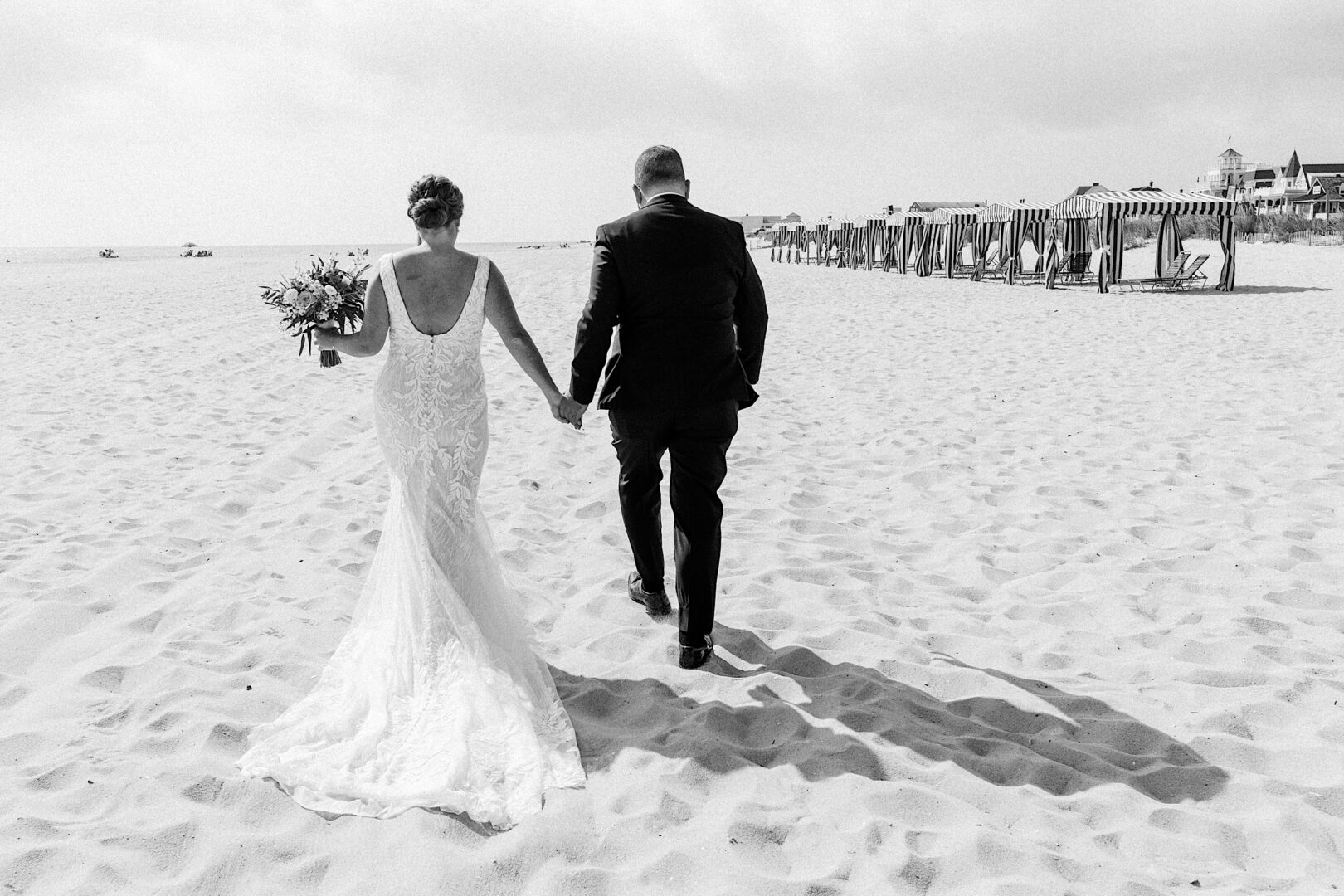 A couple in wedding attire strolls hand in hand on a sandy beach, with cabanas in the background, capturing the magic of a Corinthian Yacht Club of Cape May wedding.