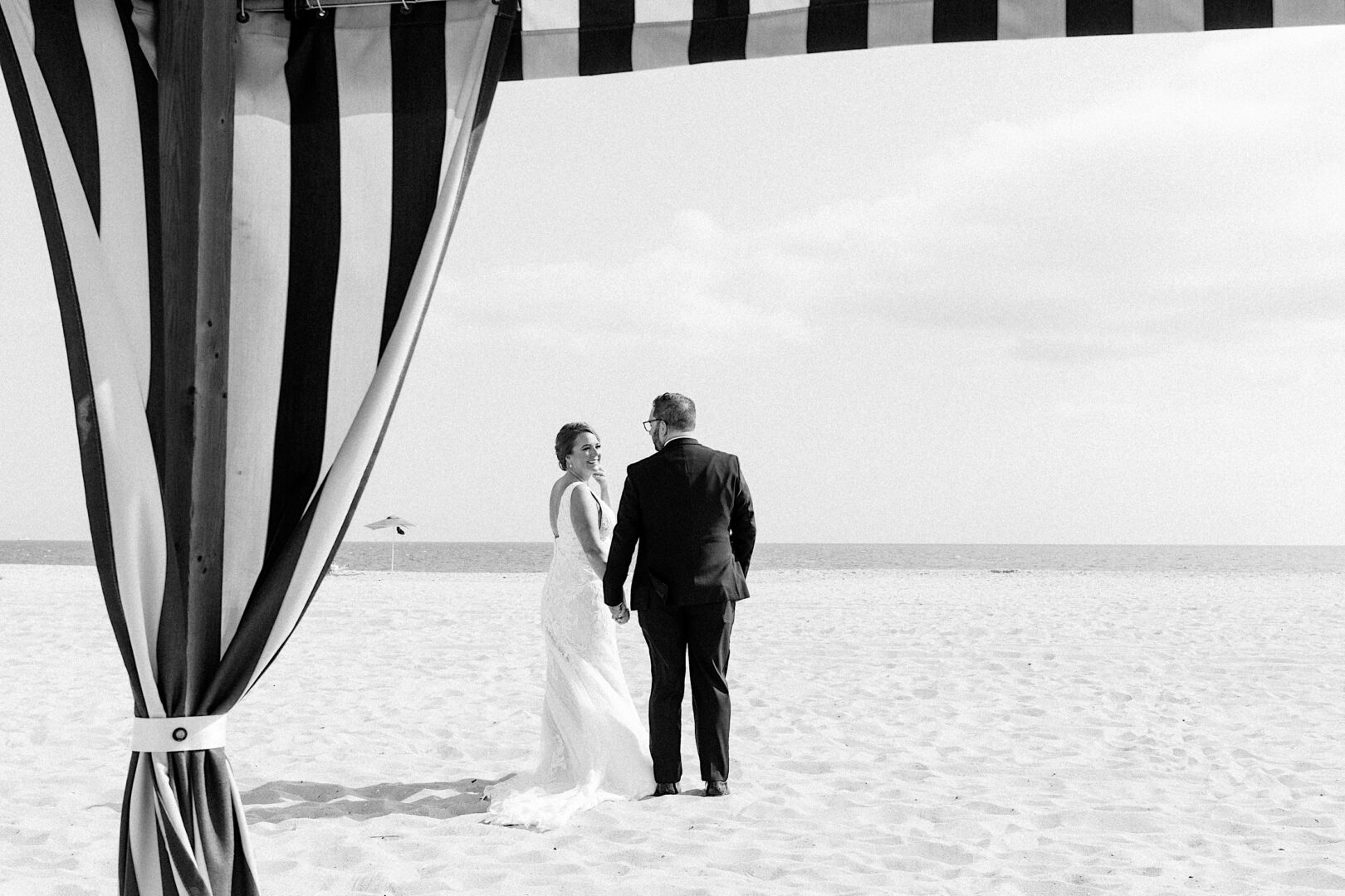 A couple in formal attire holds hands, standing on a sandy beach under a striped canopy at the Corinthian Yacht Club of Cape May, gazing towards the ocean.