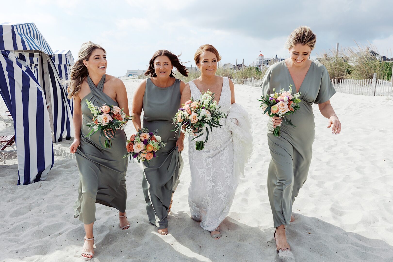 A bride in a white dress walks on the beach at the Corinthian Yacht Club of Cape May, flanked by three bridesmaids in matching sage green dresses, each holding bouquets.