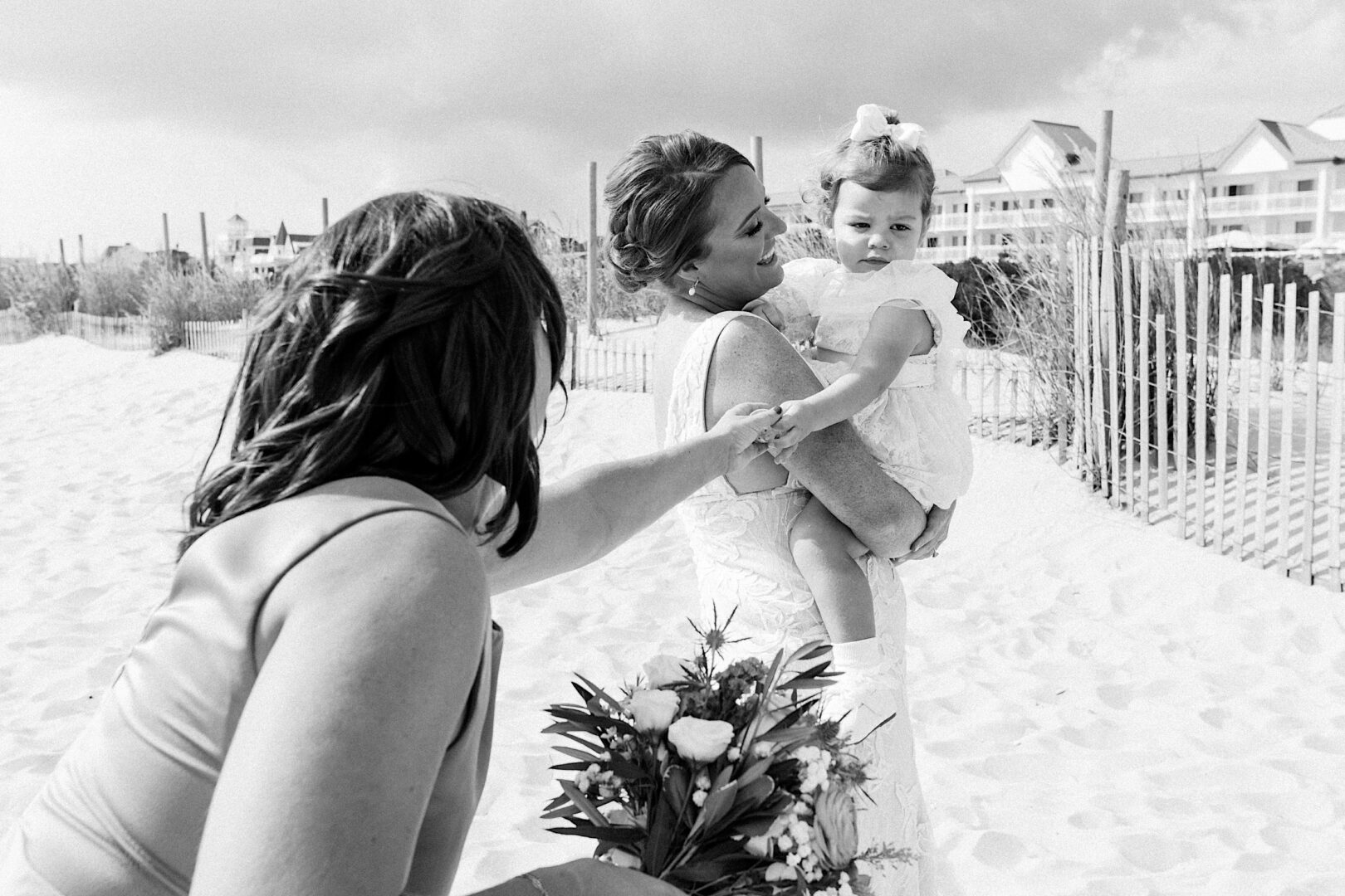 At a Corinthian Yacht Club of Cape May wedding, a woman in a stunning wedding dress cradles a child on the sandy beach, while another woman lovingly reaches out with a bouquet.