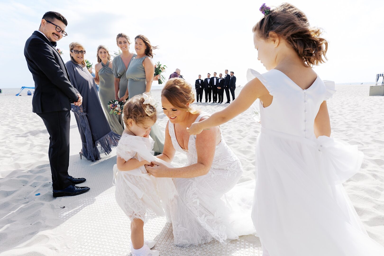 On the sandy shores near the Corinthian Yacht Club of Cape May, a bride kneels to adjust a toddler's dress, while bridesmaids and groomsmen stand poised in the background, creating a picture-perfect wedding scene.