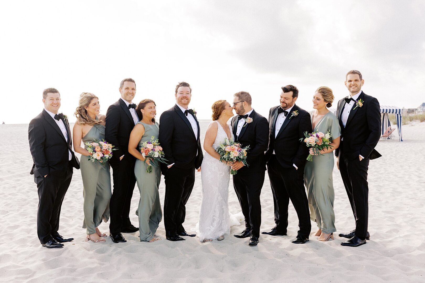 A stunning Corinthian Yacht Club of Cape May wedding unfolds on the beach, with the bride and groom radiating joy at the center. Groomsmen don black suits while bridesmaids grace elegant sage green dresses, each holding delicate bouquets in perfect harmony.