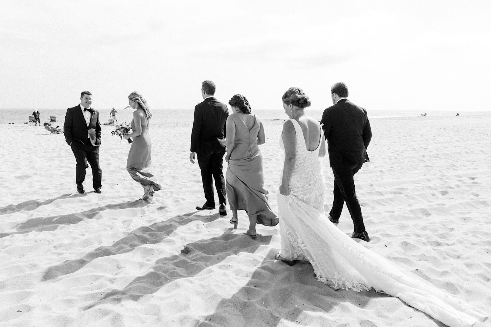 A bride and groom, adorned in formal attire, stroll with their attendants along a sandy beach, echoing the elegance of a Corinthian Yacht Club of Cape May wedding.