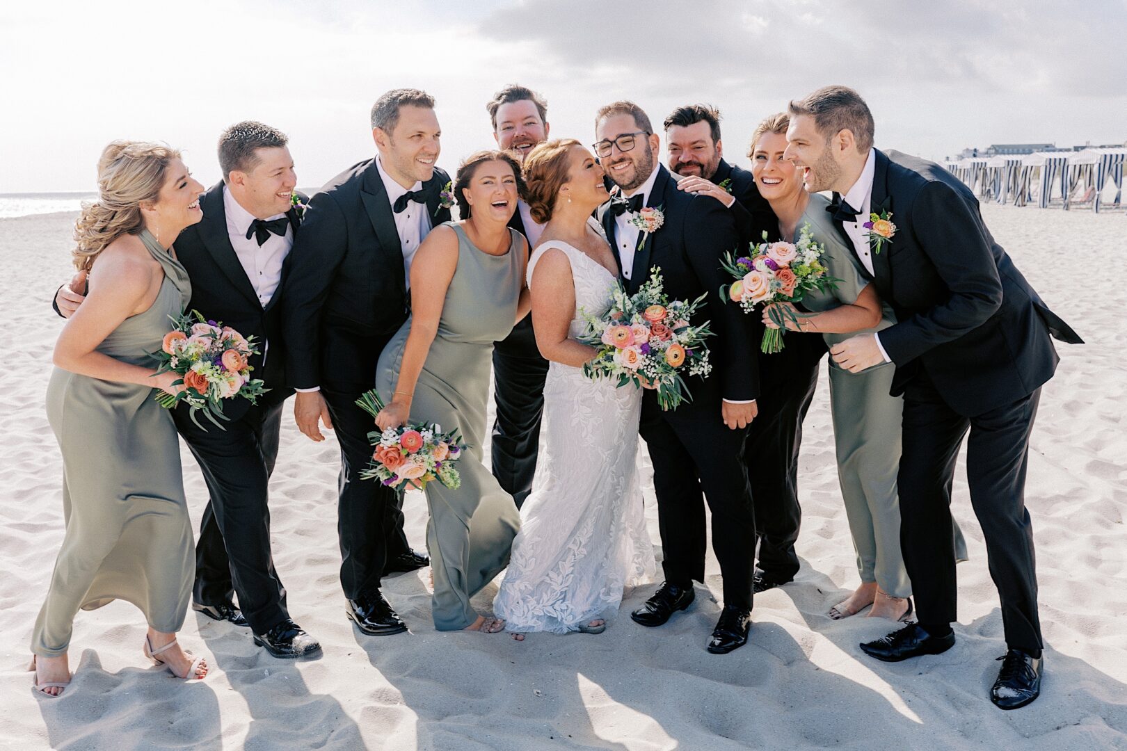 A bridal party gathers on a sandy beach at the Corinthian Yacht Club of Cape May Wedding. With the bride and groom at the center, they wear black suits and green dresses, holding floral bouquets while smiling and laughing.