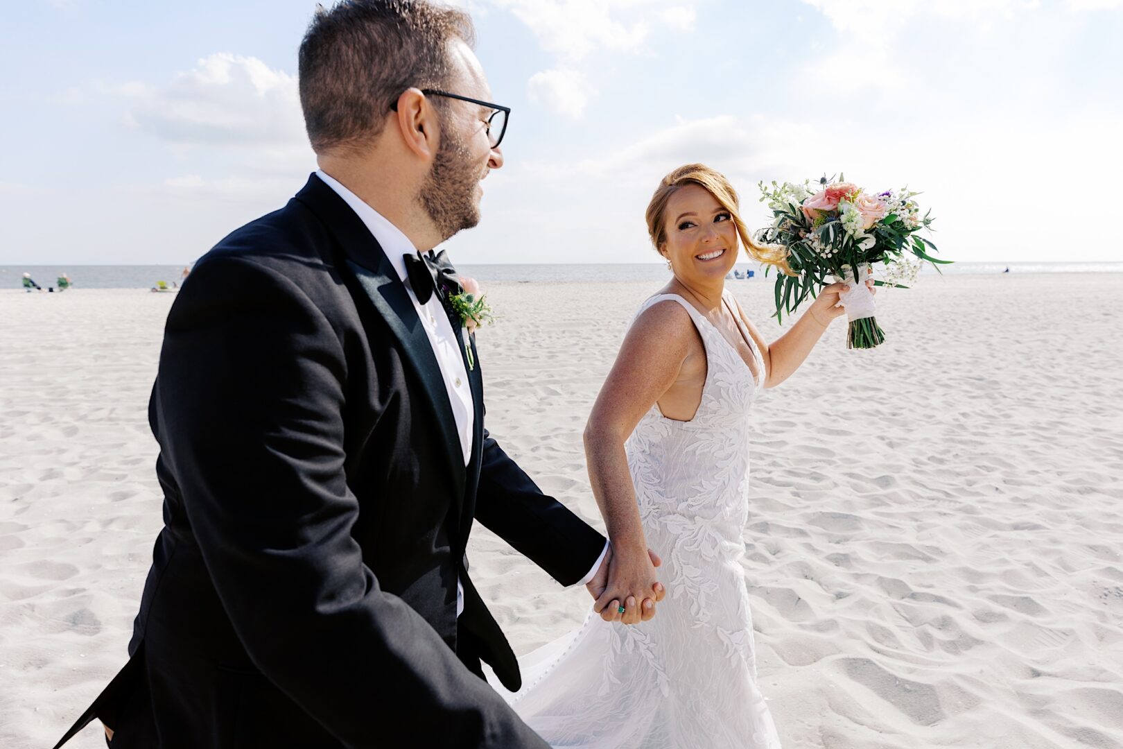 A couple, dressed in wedding attire, holds hands and smiles while walking on a sandy beach at their Corinthian Yacht Club of Cape May wedding. The bride carries a bouquet, capturing the joy of their special day.