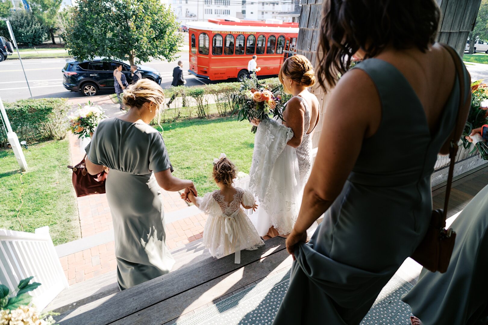 A bride and her bridesmaids, elegantly dressed in light-colored gowns, gracefully descend the steps with a young girl in white. In the background, a red trolley glides past, capturing the essence of a Corinthian Yacht Club of Cape May wedding.