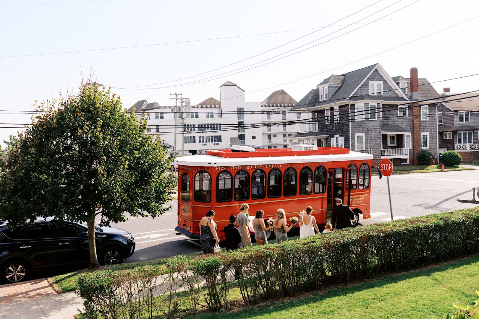 At a street corner, a red trolley bus paused, with people gathered nearby. Residential houses and a tree formed the backdrop, as if waiting to escort guests to the elegant Corinthian Yacht Club of Cape May Wedding beyond.