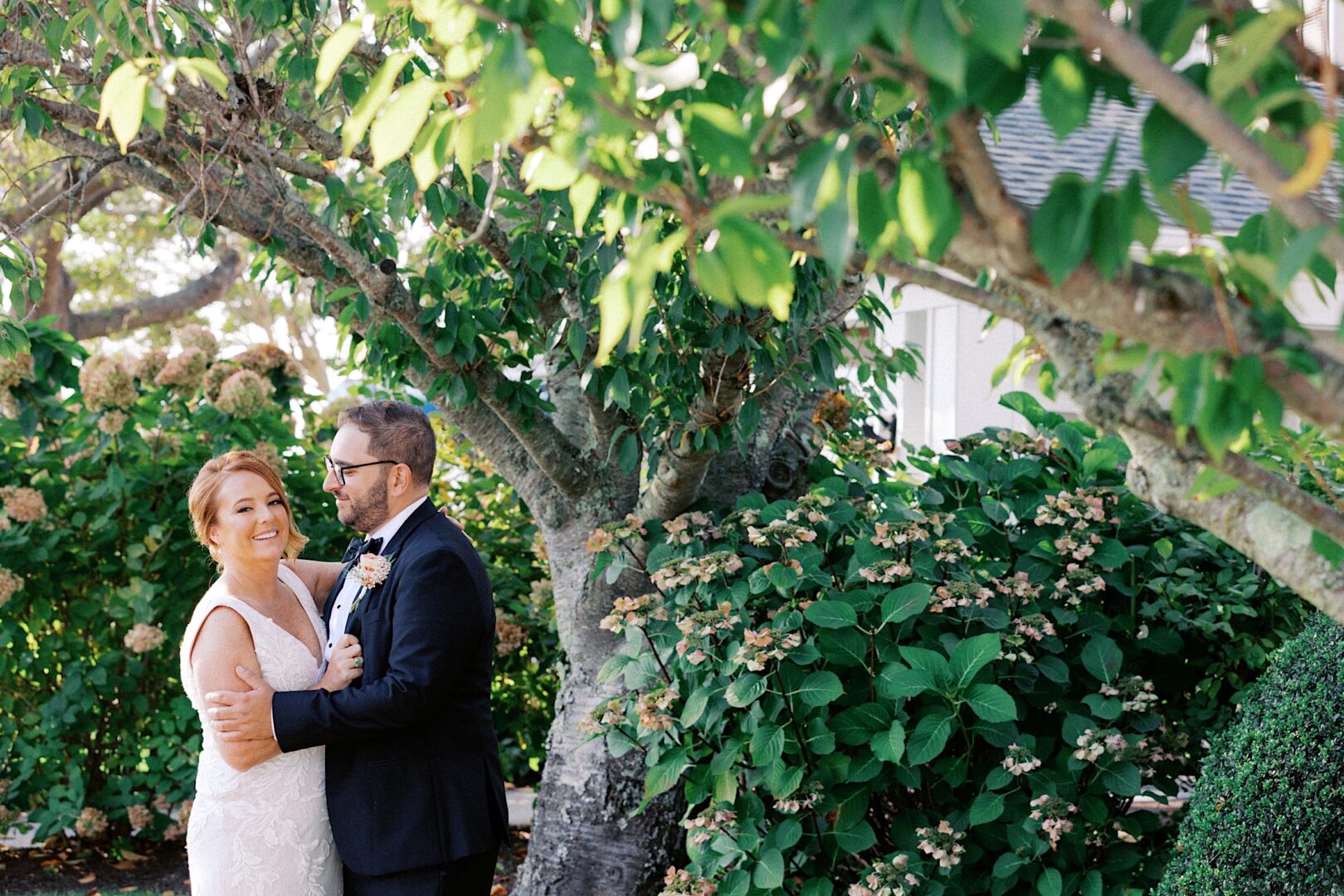 A couple in wedding attire poses under a leafy tree at the Corinthian Yacht Club of Cape May, surrounded by vibrant greenery and blossoms.