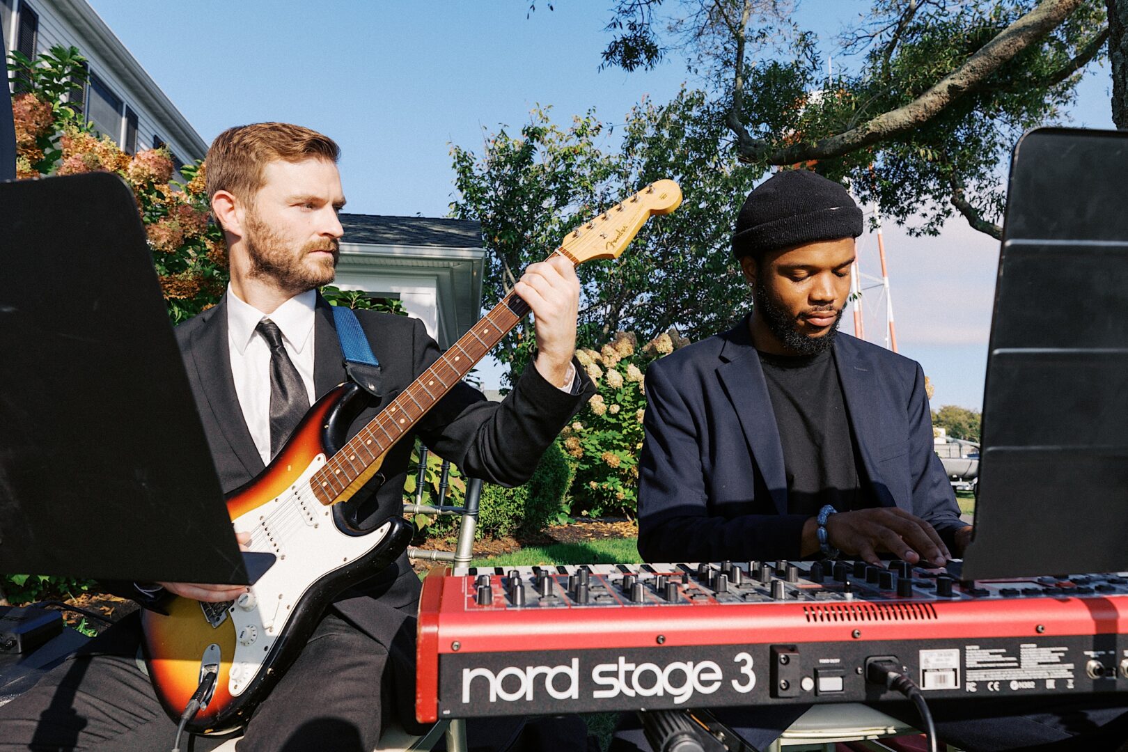 At the Corinthian Yacht Club of Cape May wedding, two musicians perform outdoors; one strums an electric guitar while the other dances across a keyboard. Dressed in dark suits and seated with grace, they captivate guests with their melodies amidst the nautical elegance.