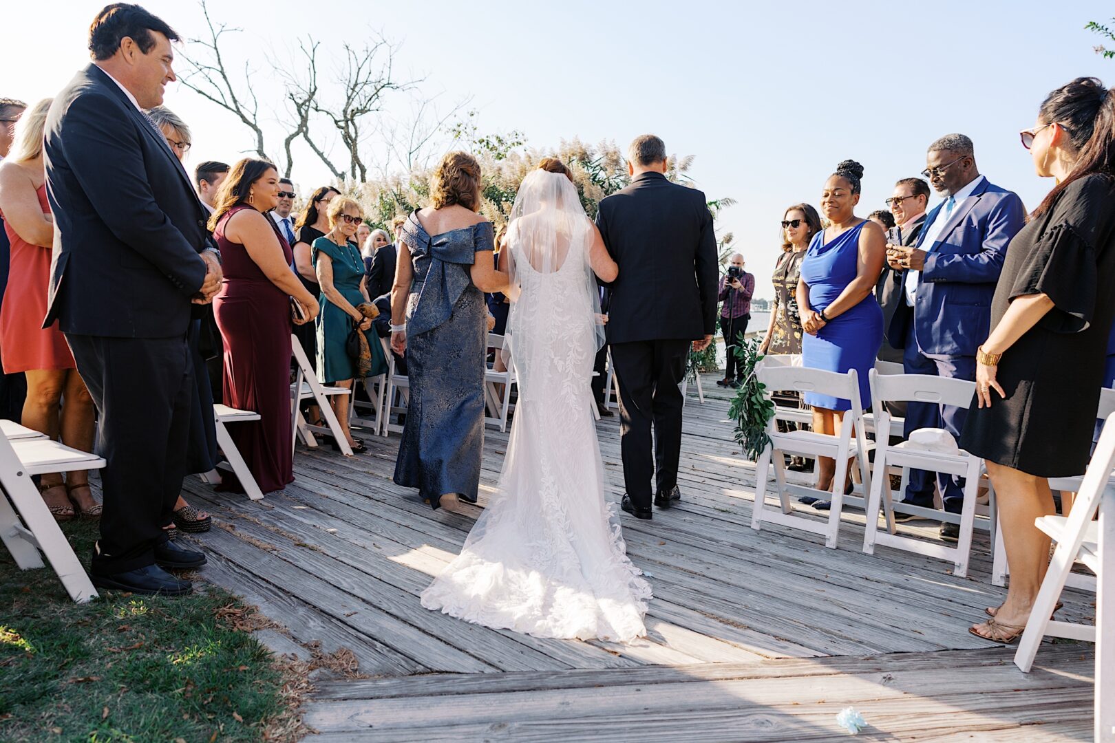 At the Corinthian Yacht Club of Cape May wedding, a bride gracefully walks down an outdoor aisle accompanied by two people. Guests are seated on white chairs, with the serene backdrop of trees enhancing the coastal elegance of the occasion.