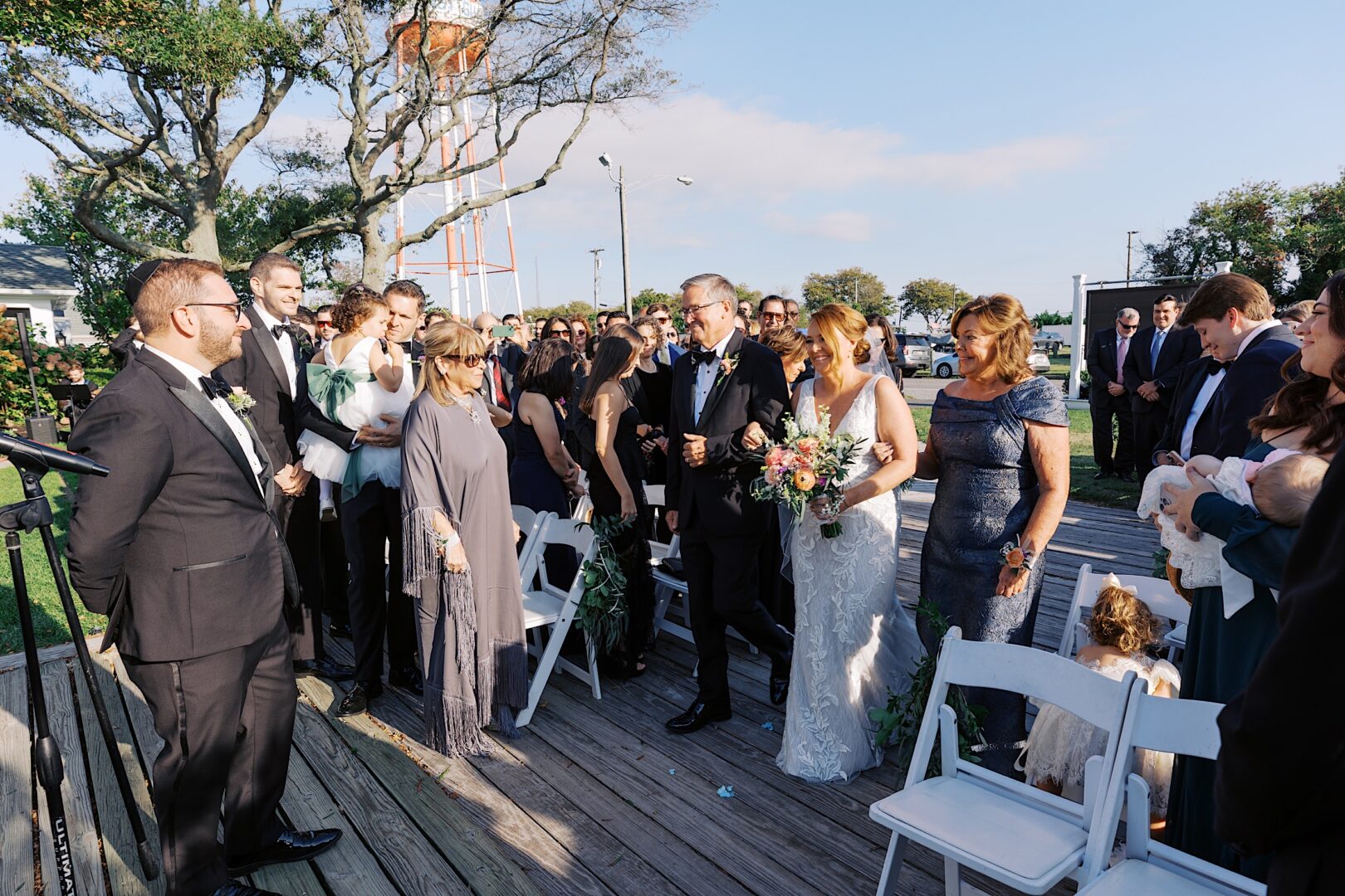 Under a clear sky, a bride walks down the aisle at the Corinthian Yacht Club of Cape May, escorted by an older man. Guests are seated on either side, and the groom eagerly awaits her at the front.