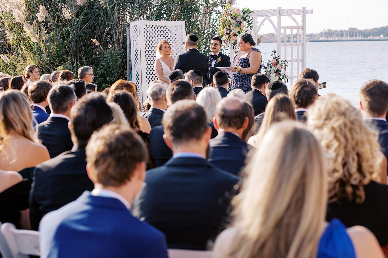 A beautiful Corinthian Yacht Club of Cape May wedding unfolds by the lakeside, where the bride, groom, and officiant stand at the altar. Guests are seated elegantly, taking in the serene atmosphere as love is celebrated.