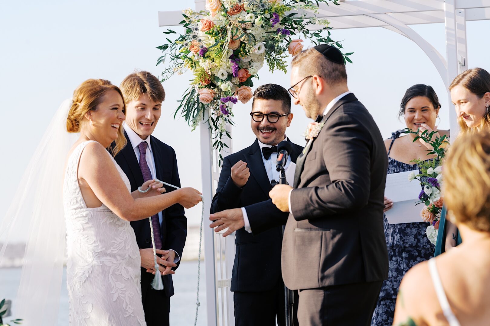 At the Corinthian Yacht Club of Cape May, a couple exchanges rings under a floral arch during their wedding ceremony, surrounded by smiling guests.