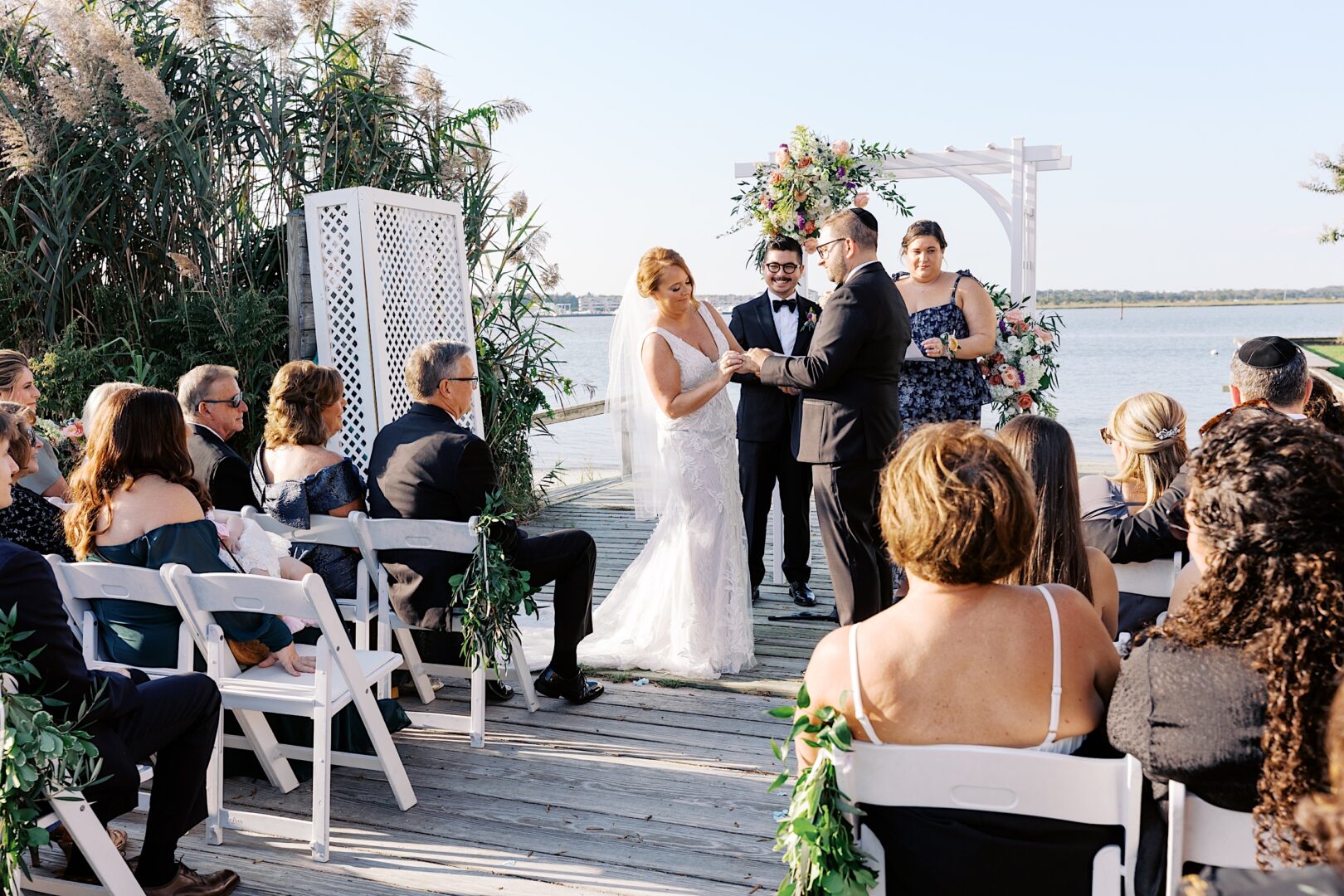 Amidst the lush greenery and seated guests, a bride and groom exchange vows by the tranquil waterfront at the elegant Corinthian Yacht Club of Cape May, creating a picture-perfect wedding moment.