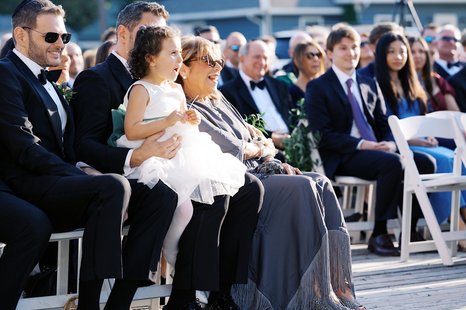 Guests seated at an outdoor Corinthian Yacht Club of Cape May wedding, including a man holding a young girl in a white dress. Others around them are smiling and dressed in formal attire.