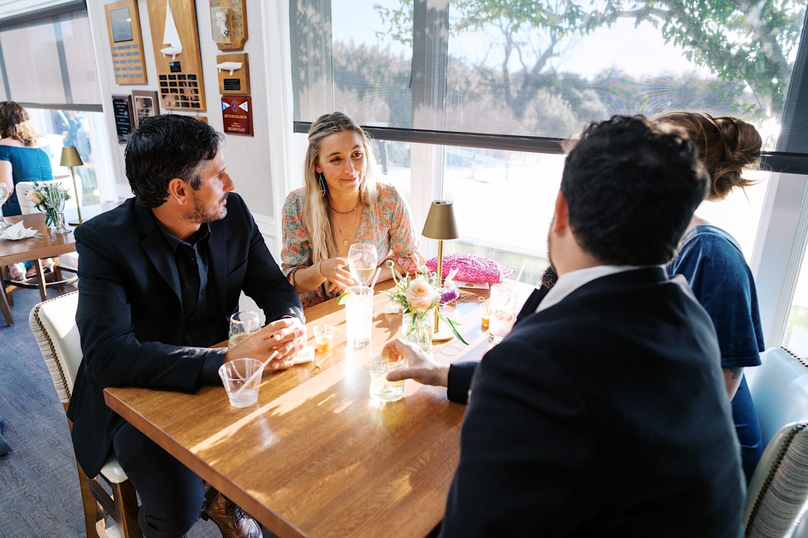 Four people in formal attire gather around a wooden table adorned with drinks and a flower vase, their faces bathed in the warm, sunlit glow of the Corinthian Yacht Club of Cape May wedding setting.